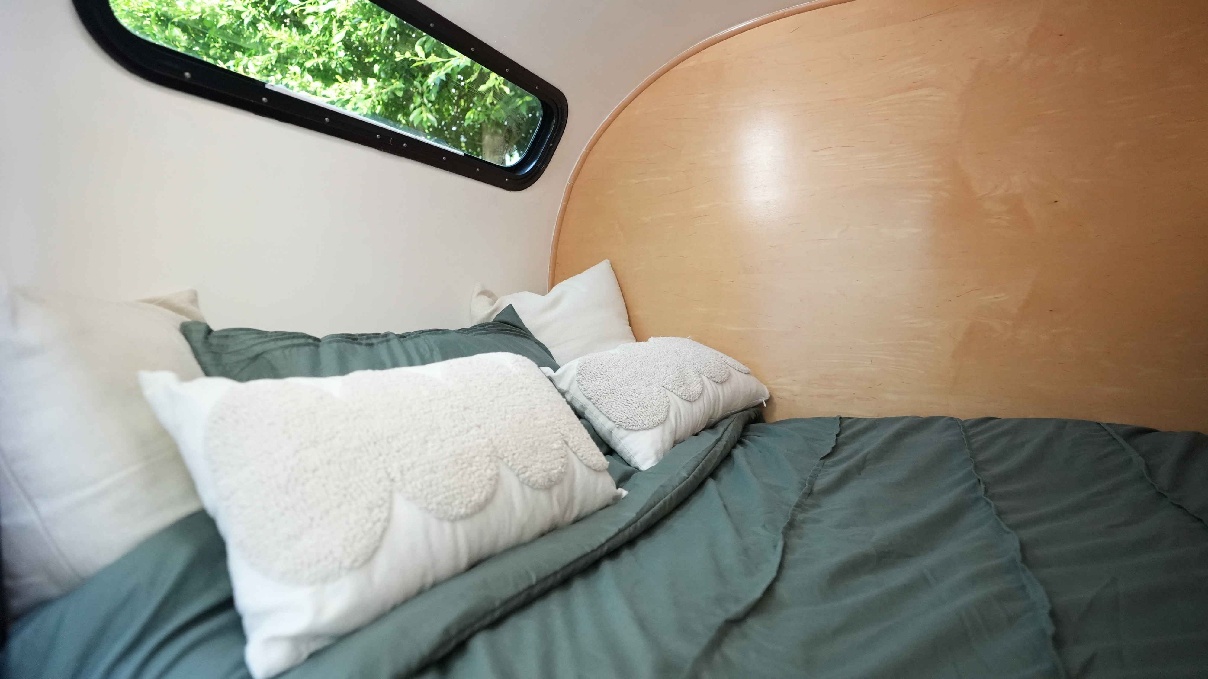 Inside this cozy rv rental: a comfy bed with green bedding, white scalloped pillows, and a window view of lush trees.. Long Teardrop 2005