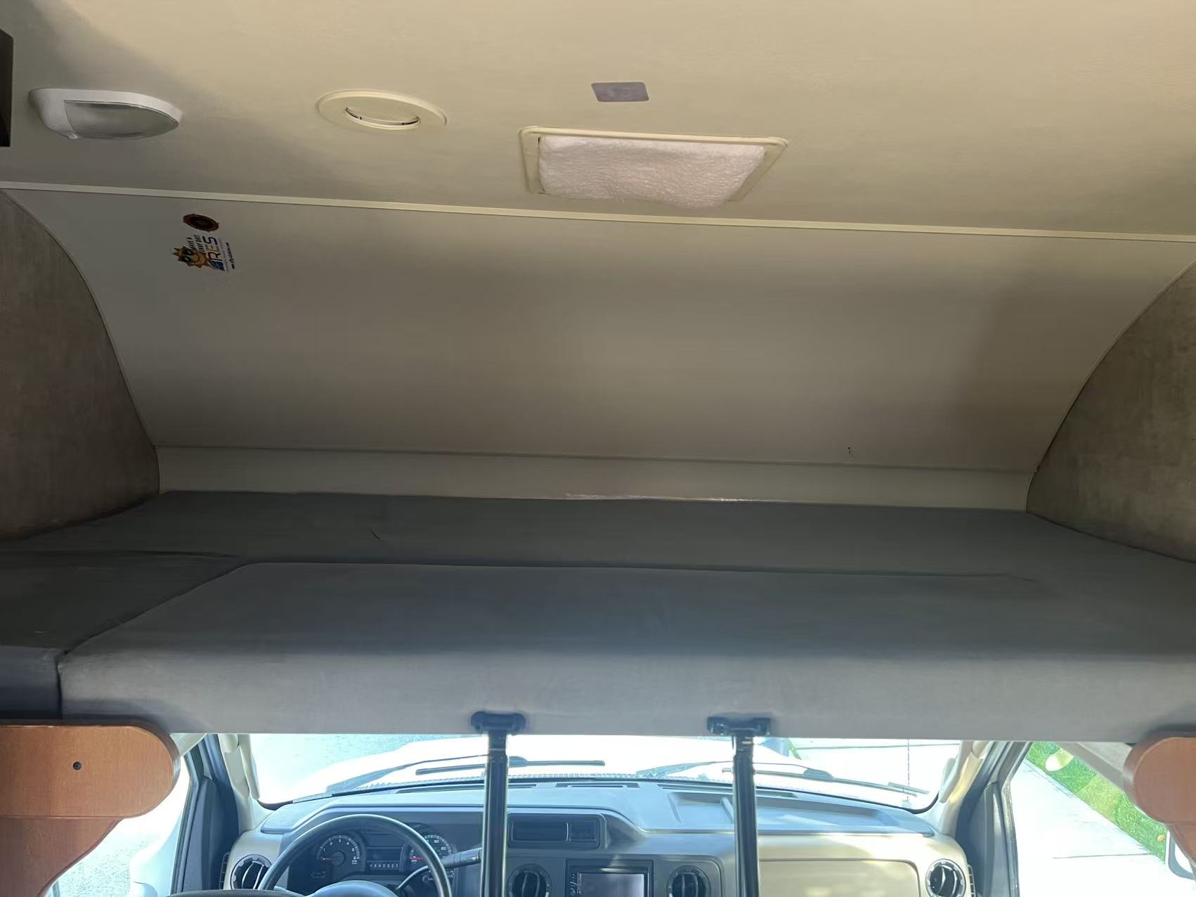 Inside an RV rental, showing the cab-over bunk bed with grey mattress, overlooking the driver's dashboard.. Itasca Spirit 2014