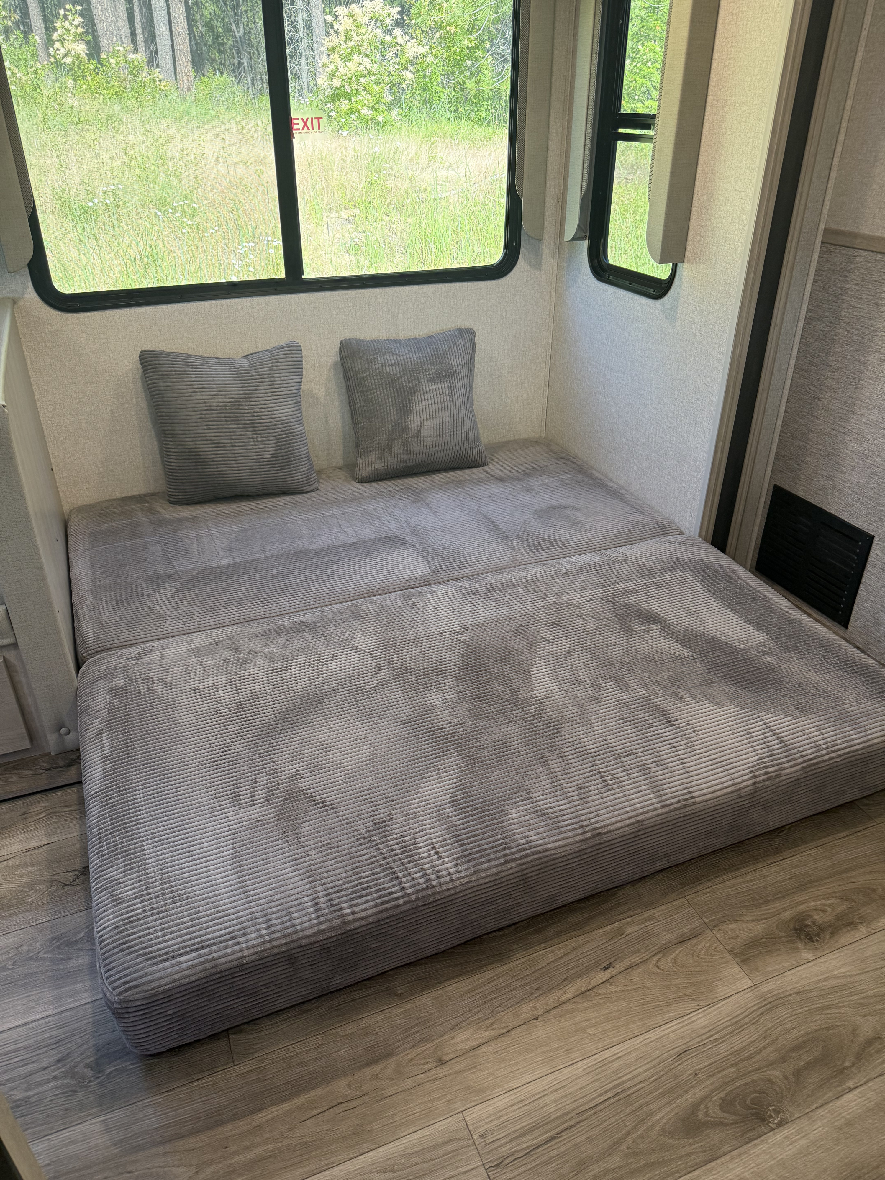 Inside an RV rental: a gray corduroy bed with pillows and a window view of green trees and grass. An EXIT sign is visible.. Forest River 2810KIK 2022