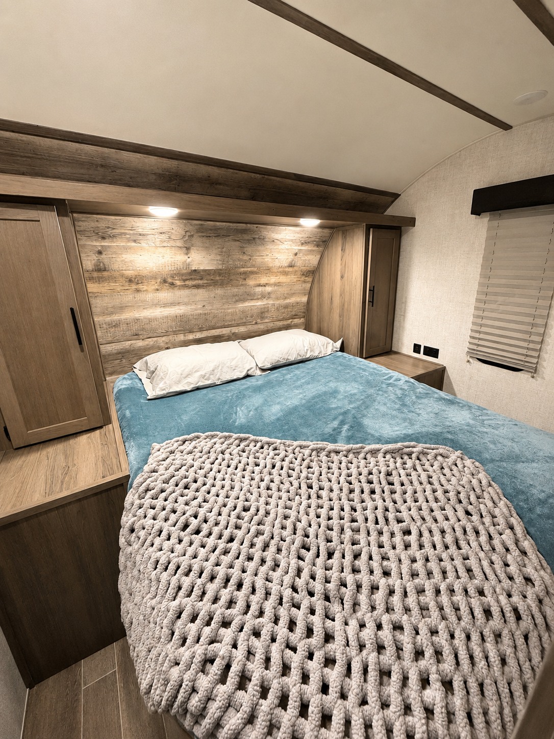 Cozy RV rental bedroom with a bed, blue bedding, white pillows, and a chunky grey knit throw. Features a wooden headboard and cabinets.. Gulf Stream Ameri-Lite 2021