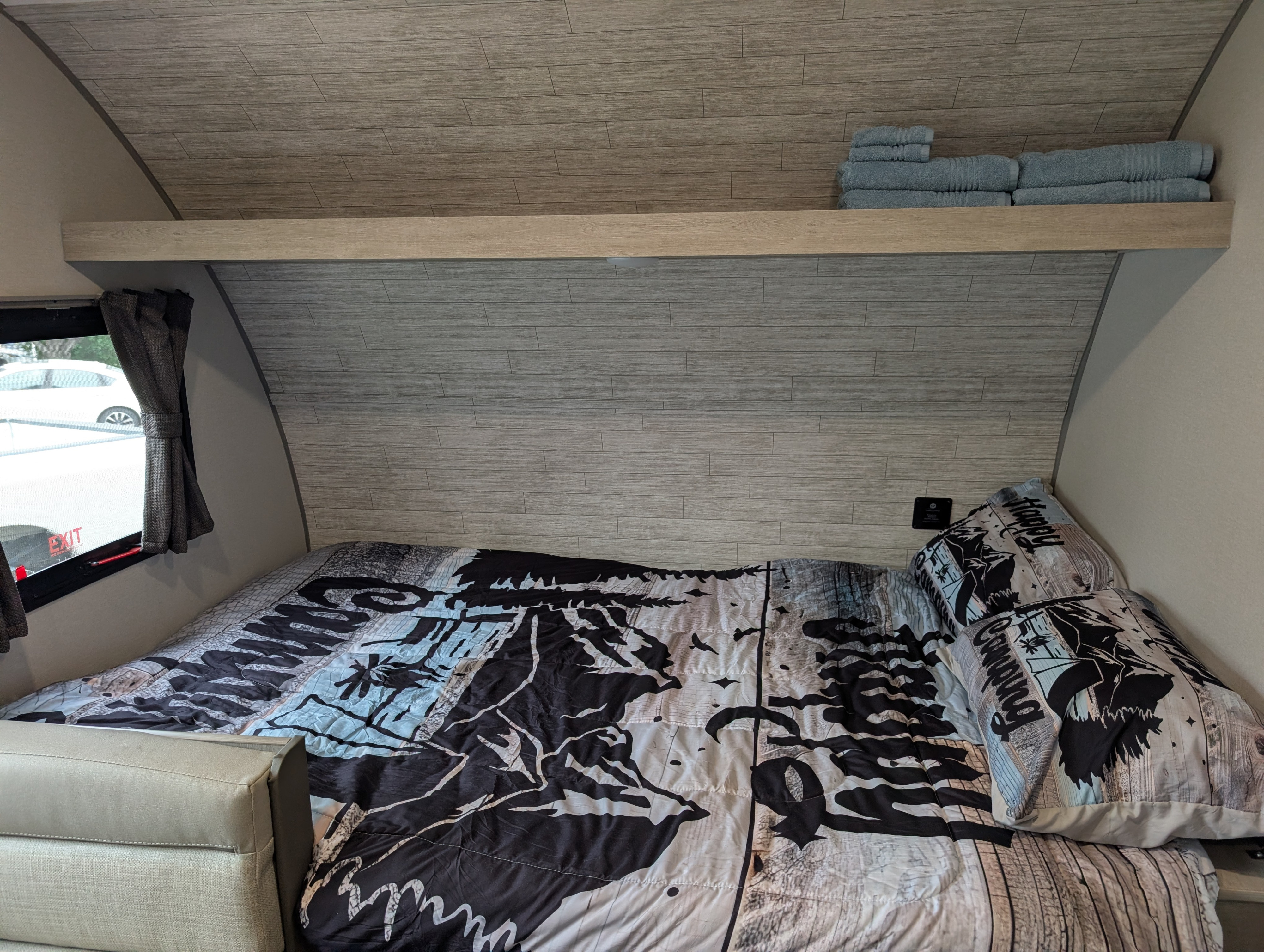 Cozy RV rental bedroom featuring wood-look walls, patterned bedding, an overhead shelf with towels, and a window.. Coachman Catilina 16bhx 2026