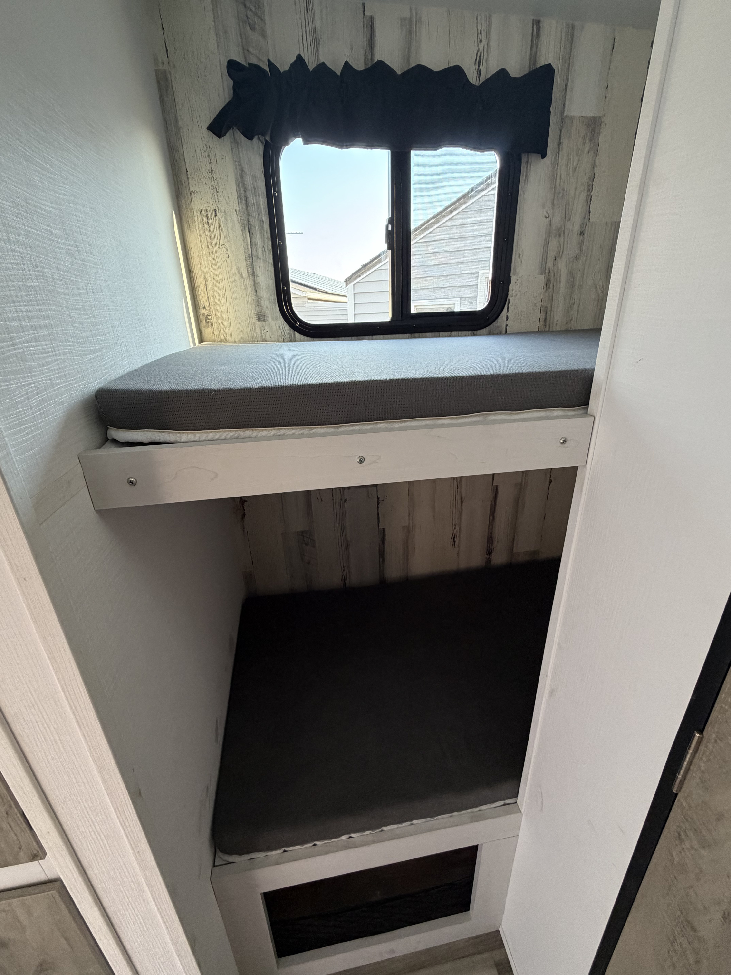 Compact bunk beds inside an RV rental. Grey mattresses on two bunks, with a window and black valance above the top bunk.. Travel Lite Rove XL 2024