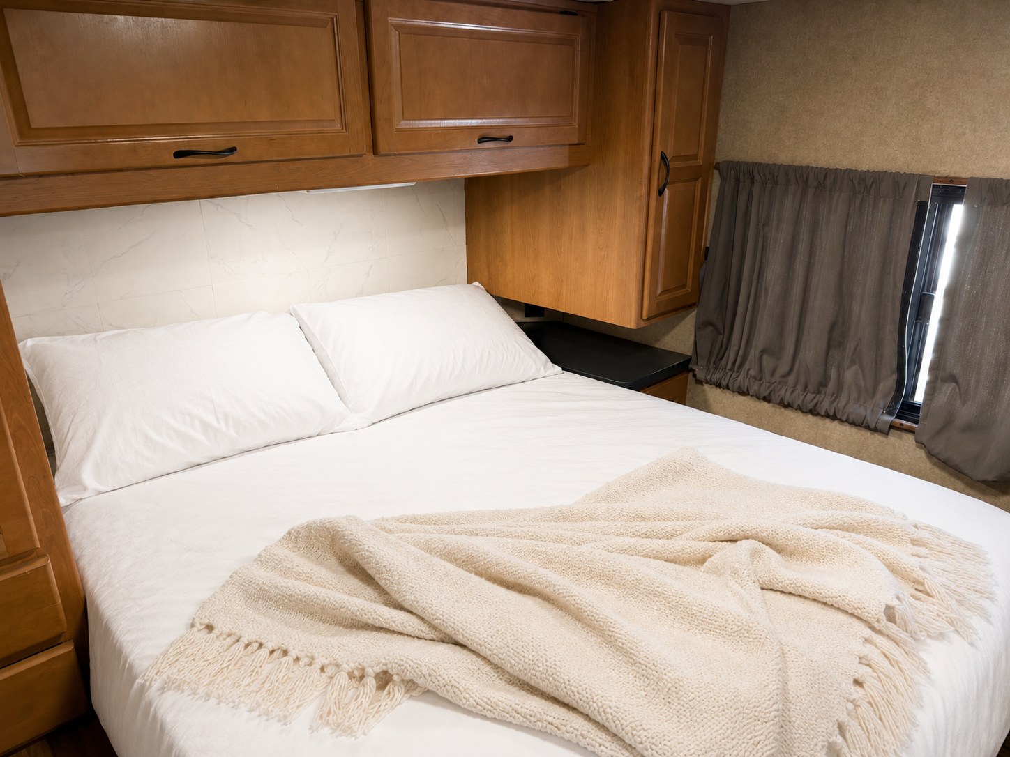 This RV rental features a cozy bedroom with a white bed, neutral blanket, overhead cabinets, and window with curtains.. Thor Majestic 2017