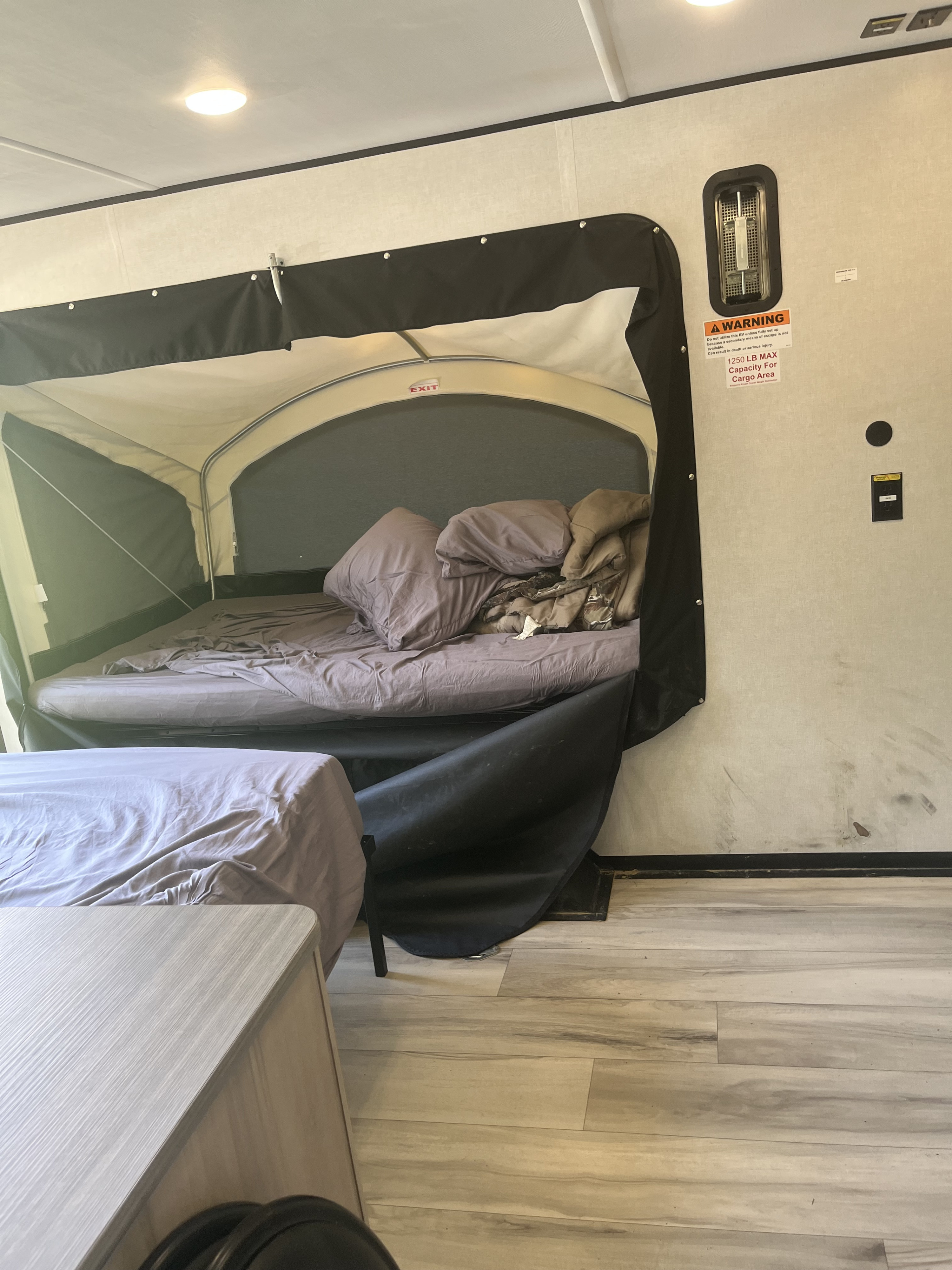 RV rental interior: A pop-up camper bed with bedding and an EXIT sign. A warning label is on the wall, and another bed sits below.. Keystone Coleman 13T 2026