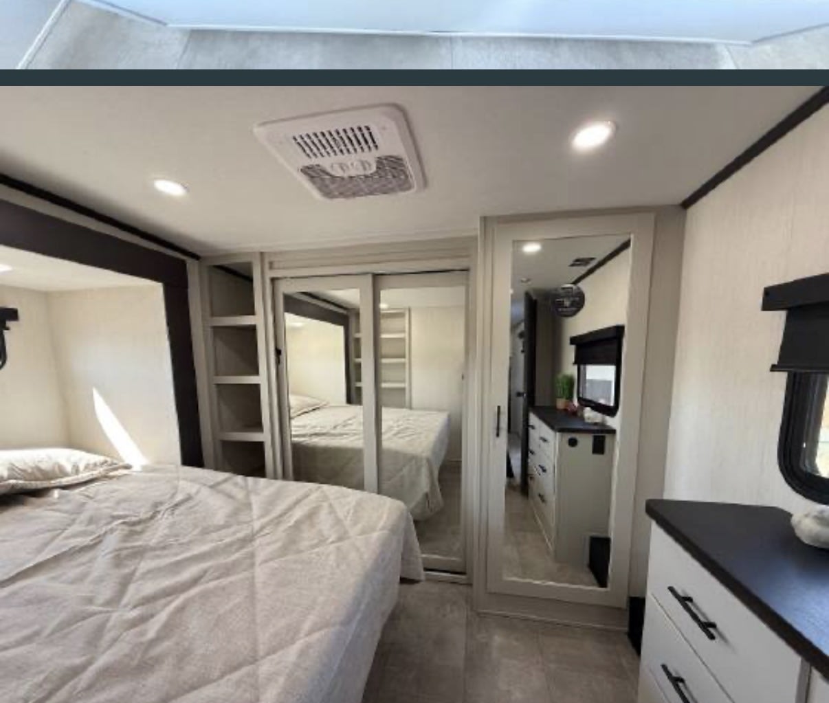 Modern RV rental bedroom with a comfortable bed, mirrored closet doors, shelves, and dresser storage. Ceiling AC unit visible.. Prime Time Rv Bonus 38 2027