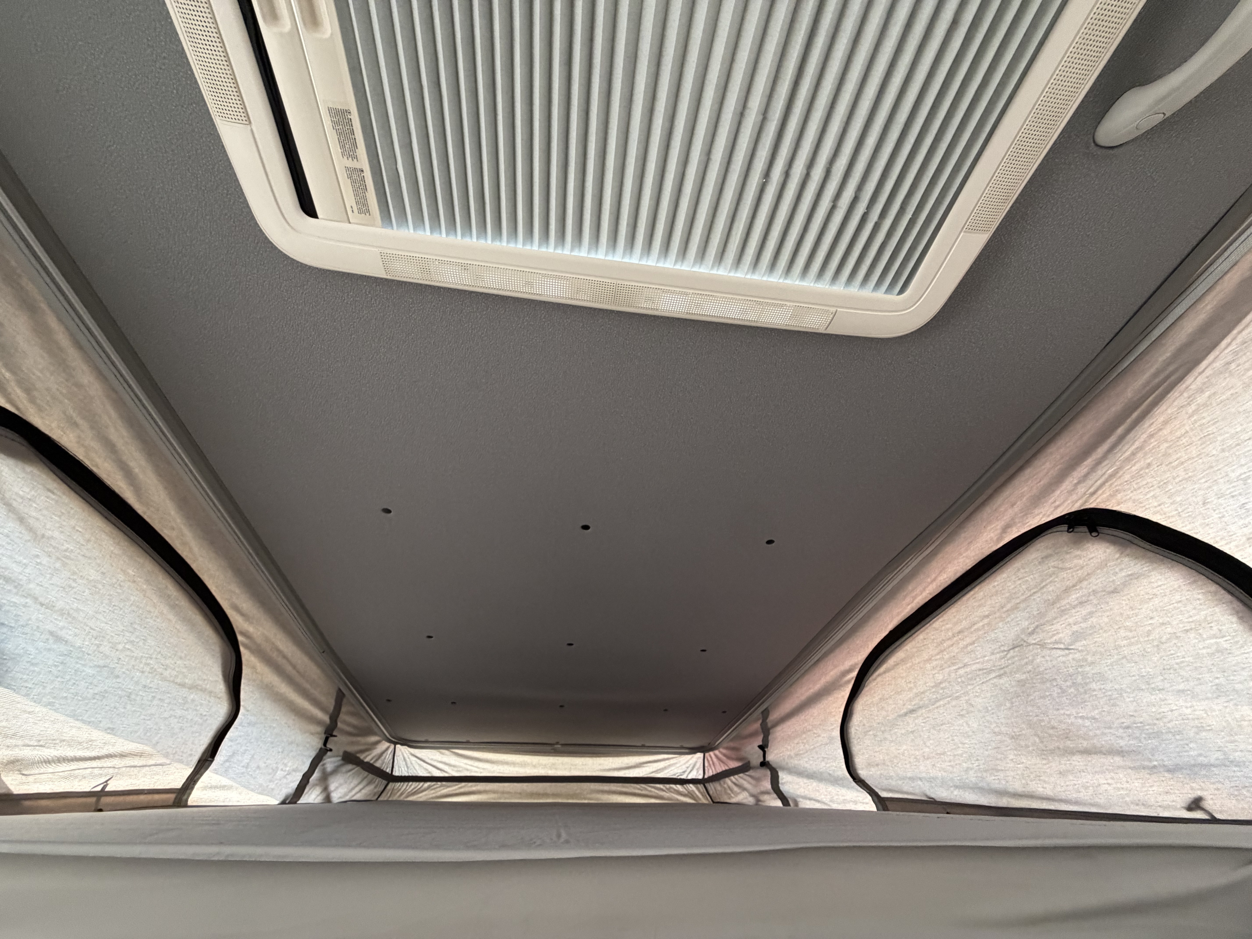 RV rental pop-top camper interior looking up at the gray ceiling with vent, pleated blinds, and upper bed.. Dodge Ram 1500 2023