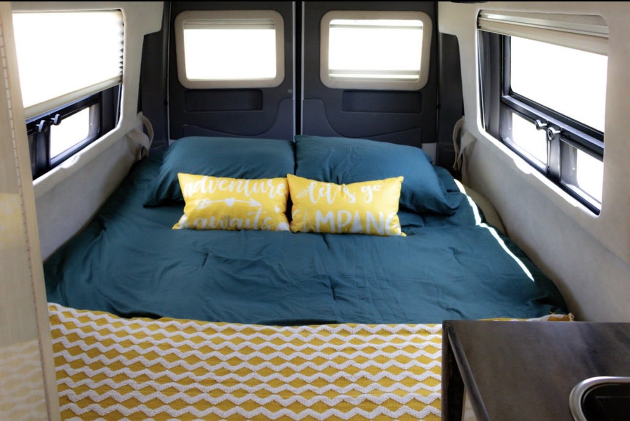 Cozy RV rental bedroom with teal bedding, yellow "adventure awaits" pillows, a chevron blanket, and bright windows.. Airstream Interstate 2014