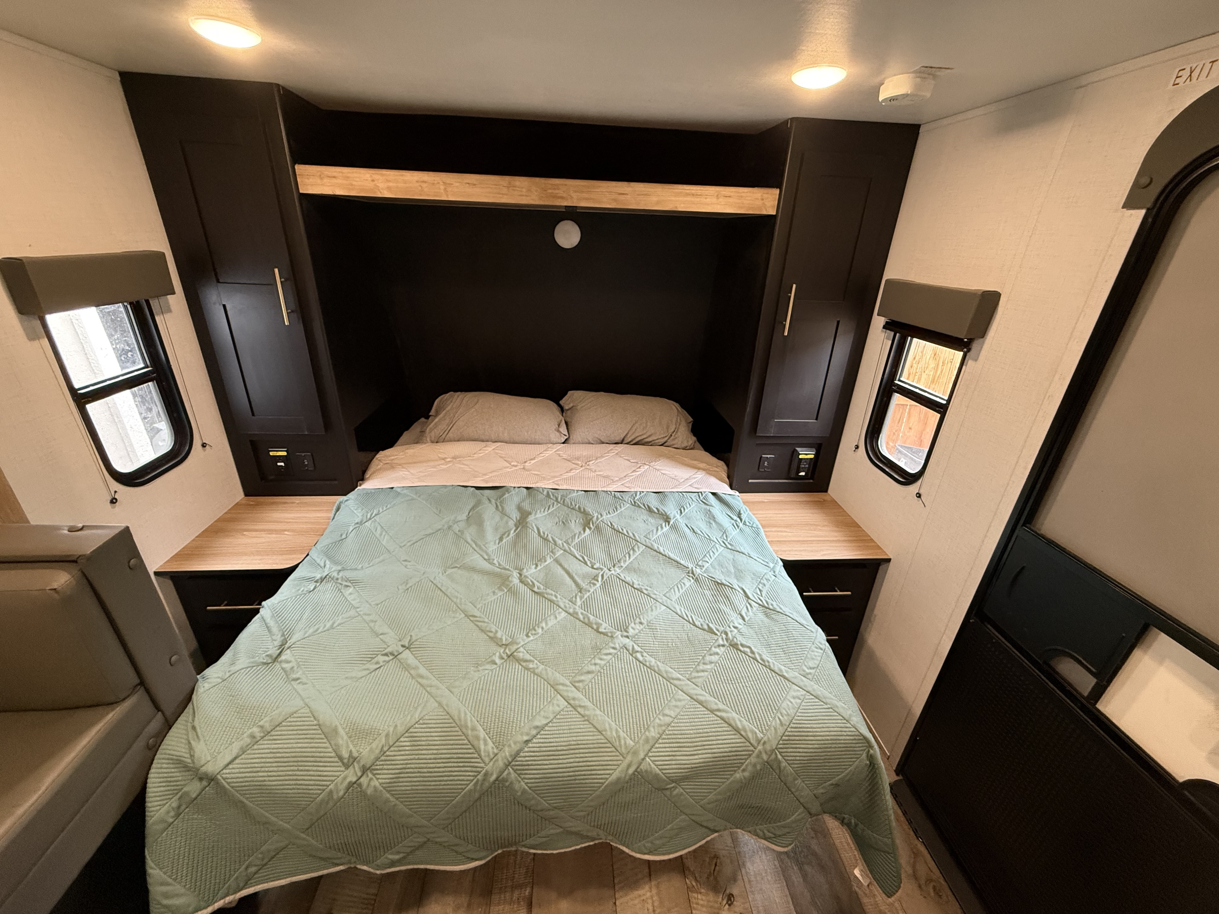 Cozy RV rental bedroom featuring a green quilted bed, dark cabinets, wooden nightstands, and windows.. Keystone RV Springdale 2023