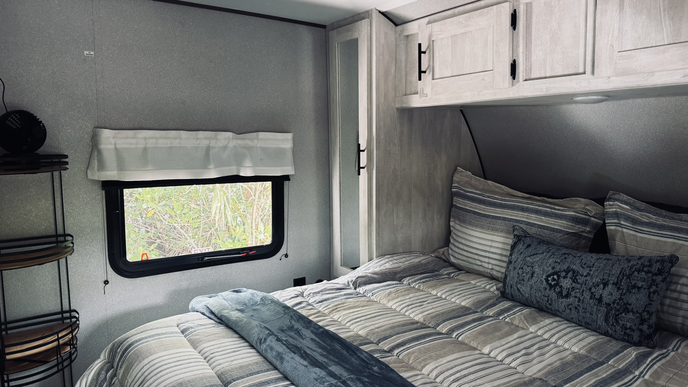 Cozy RV rental bedroom featuring striped bedding, blue throw, light cabinets, and a window to nature. Perfect for travel.. Forest River Shasta 2022