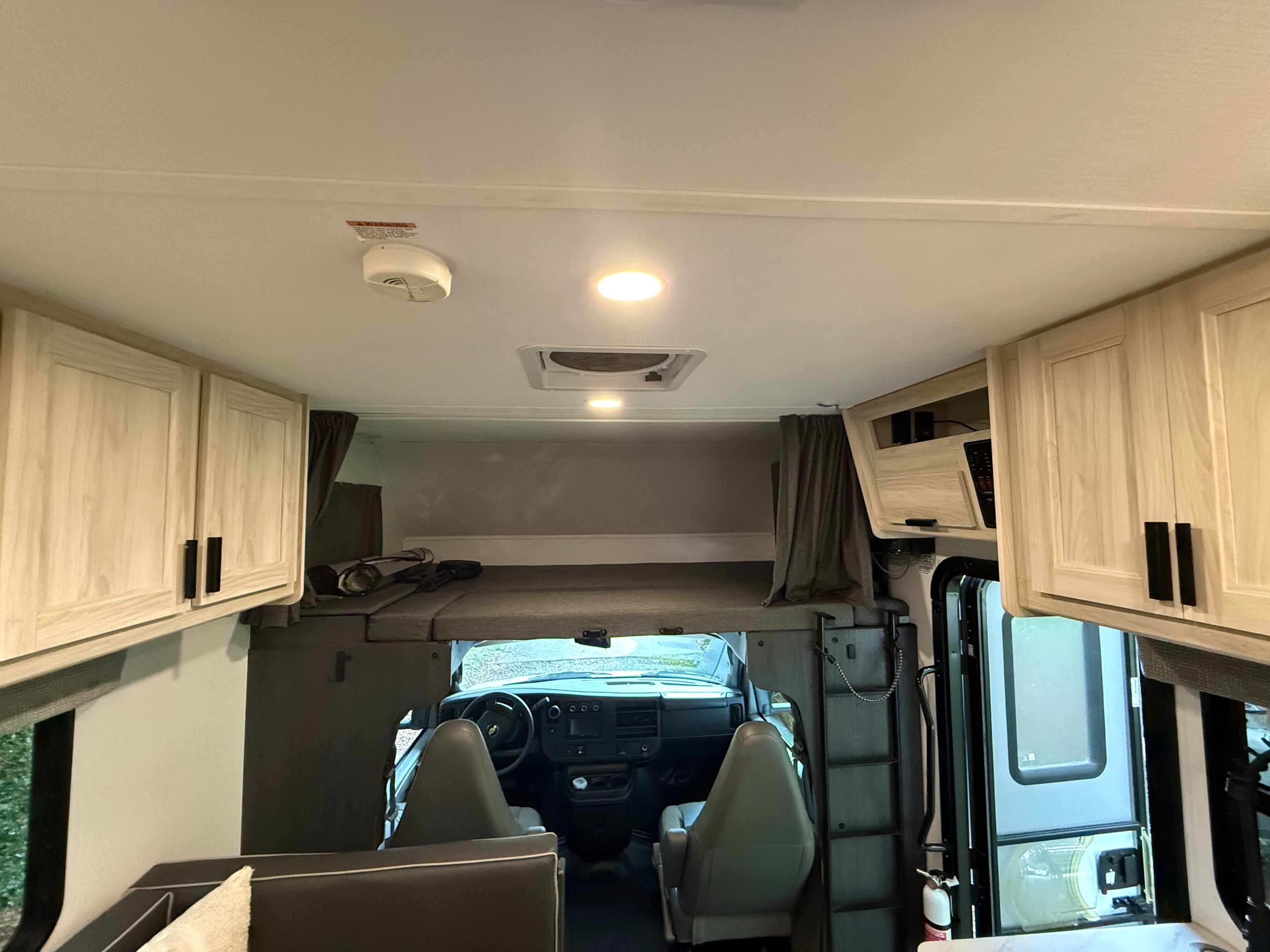 View into an rv rental's cab-over bunk and cockpit. Light wood cabinets, a vent, and a smoke detector are visible inside.. Forest River Sunseeker LE 2023