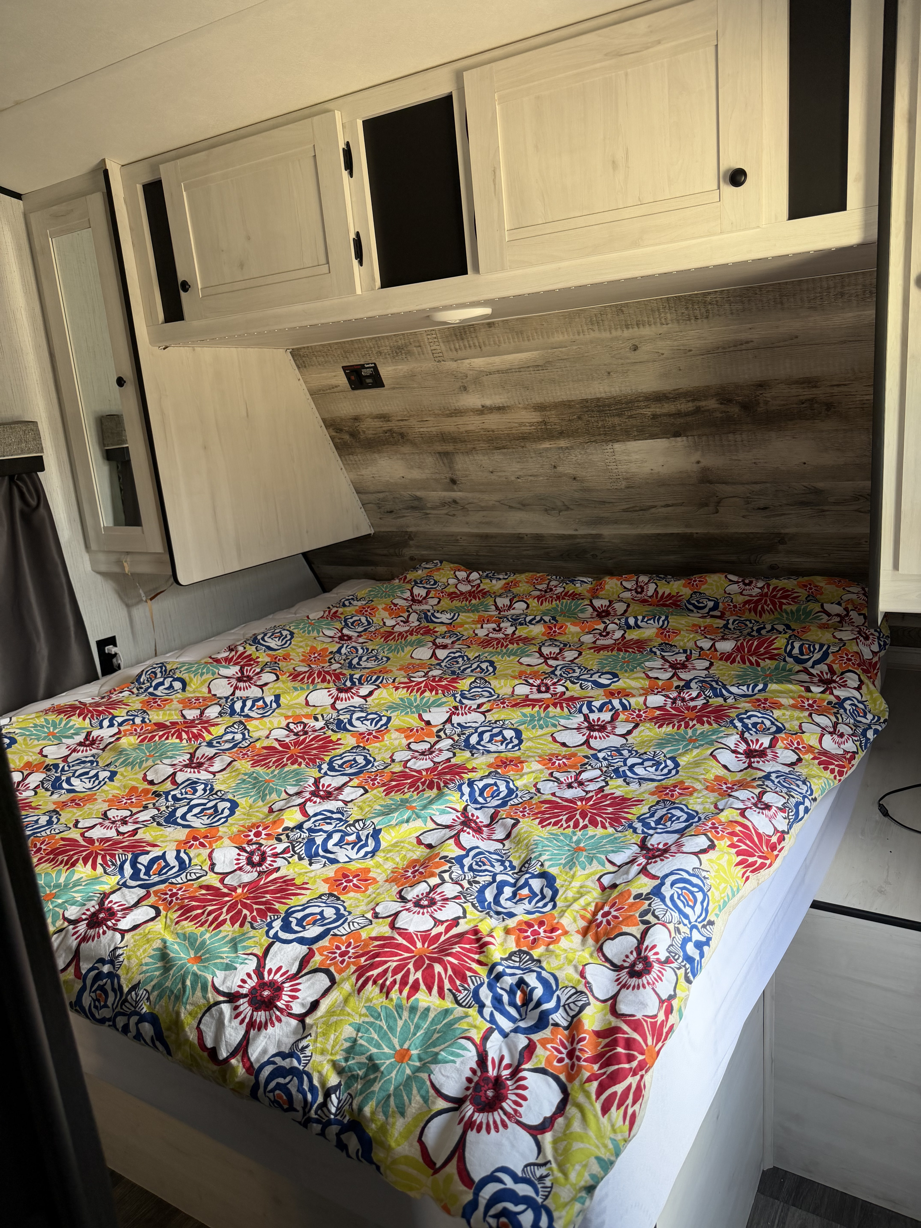 Cozy RV rental bedroom with colorful floral bedding, open overhead cabinet, and rustic wood headboard paneling.. Heartland Lithium 2021