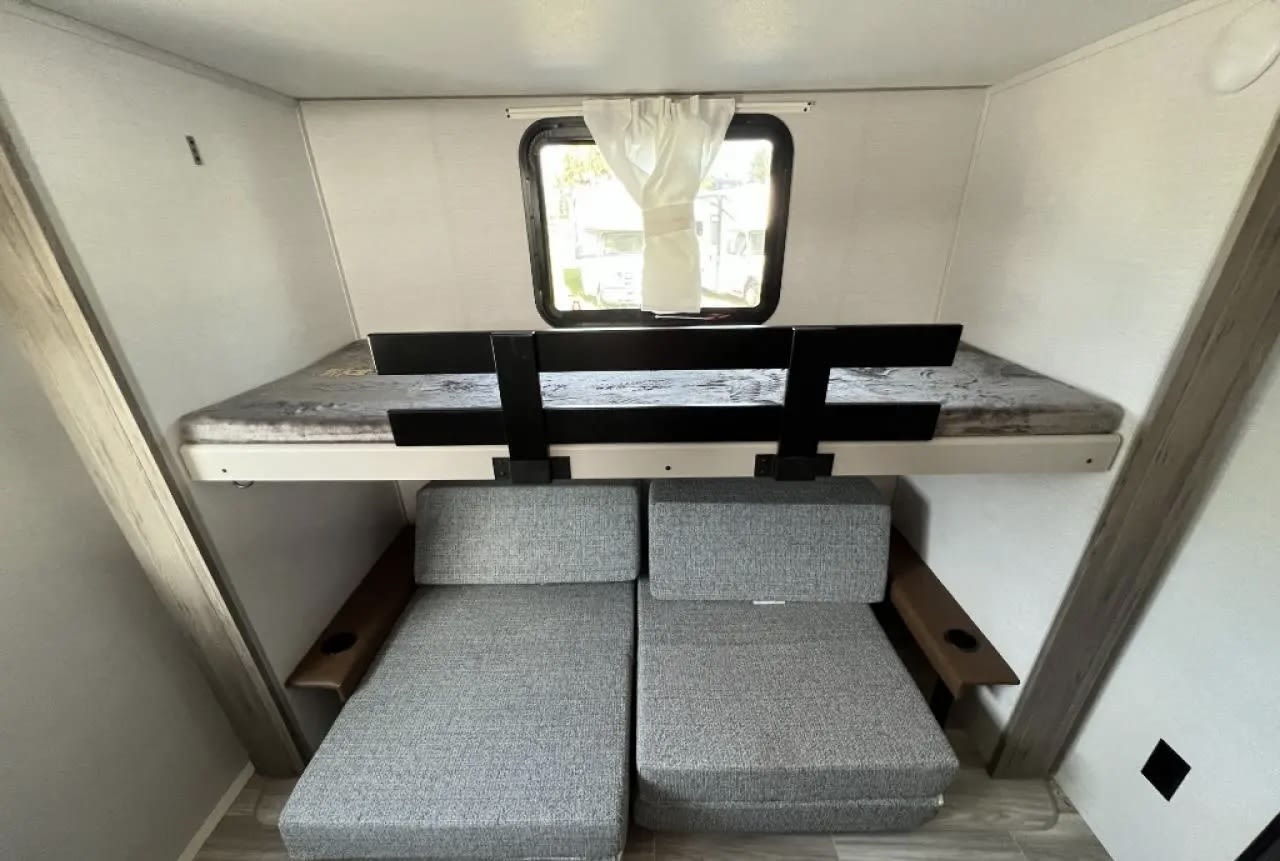 RV rental interior: Bunk bed with safety rail over two gray modular seats with armrests and cup holders, beneath a window.. Forest River Vibe 2024
