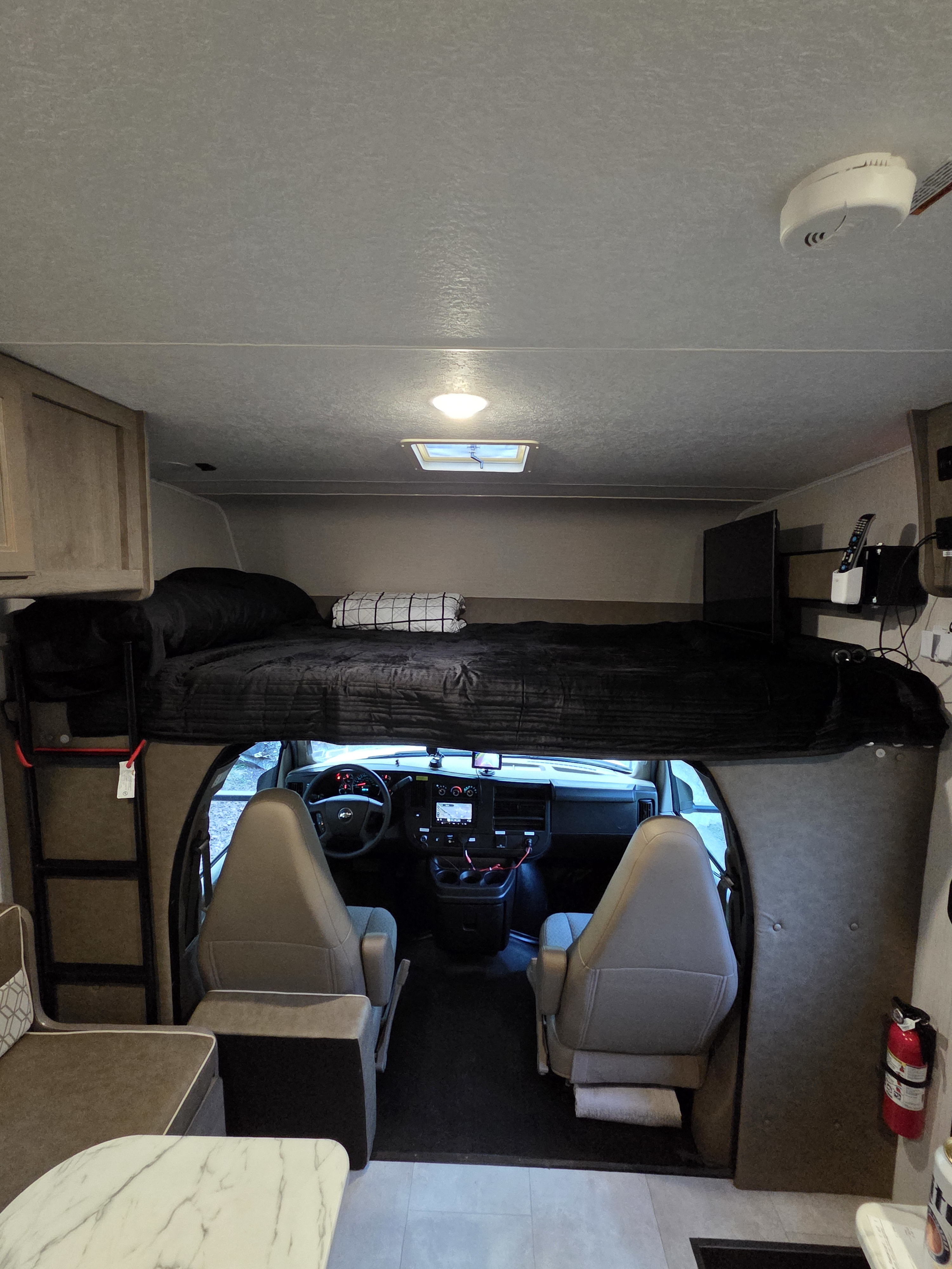 Interior of an RV rental: looking forward to the cockpit below the overhead bunk, with a ladder and dinette visible.. Forest River Coachmen Freelander 2020