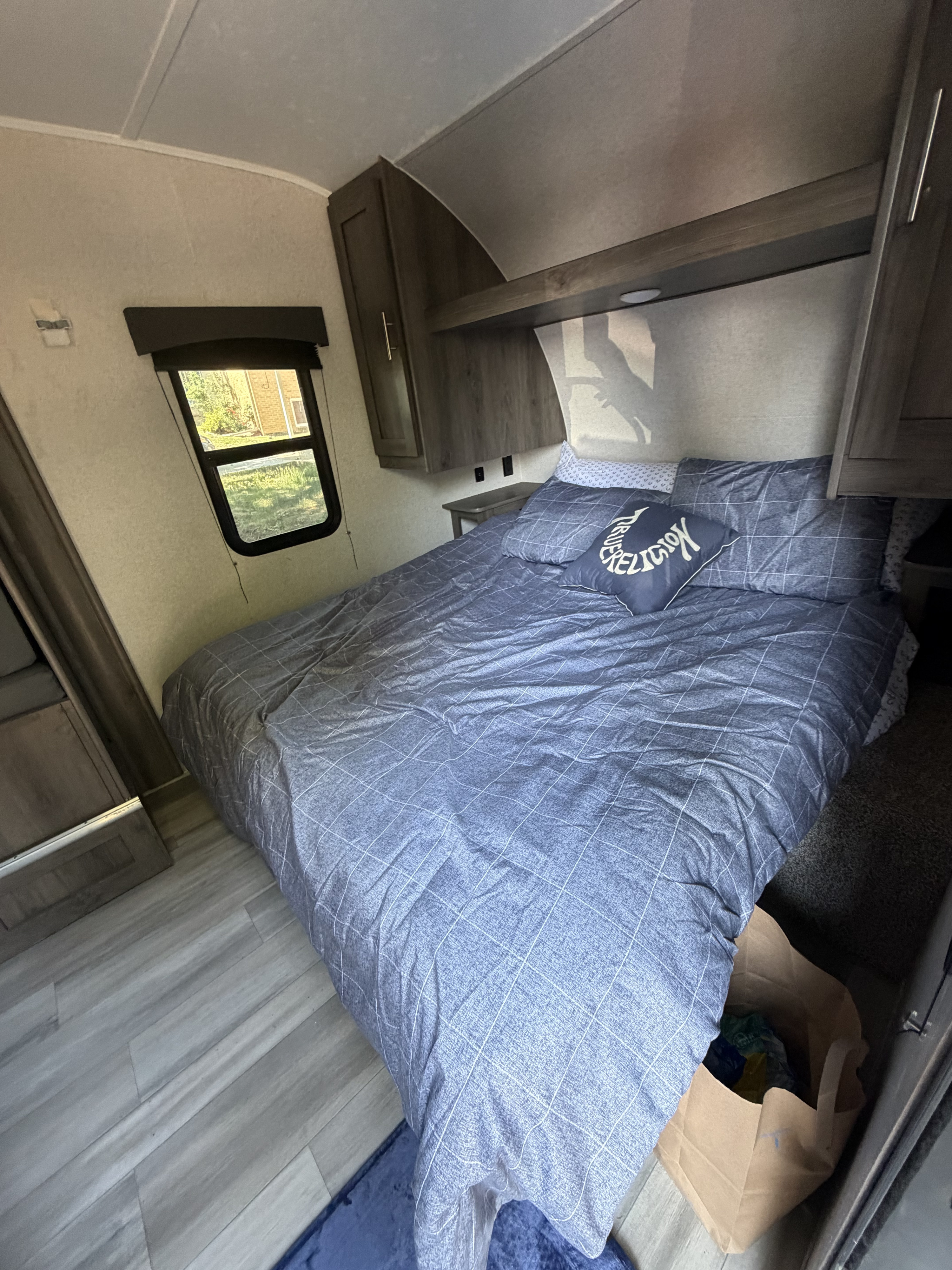 Inside an RV rental: a cozy bedroom with a blue patterned bed, overhead cabinets, and a window to nature.. Coachmen viking 2021