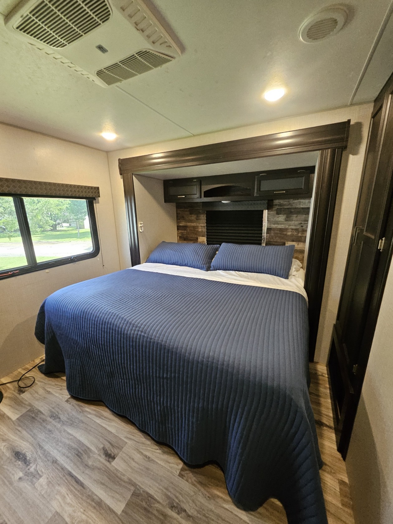 This cozy RV rental bedroom features a neatly made bed with blue bedding, a window overlooking greenery, wooden floor, and overhead cabinets.. Venture SportTrek Touring STT333VFK 2020