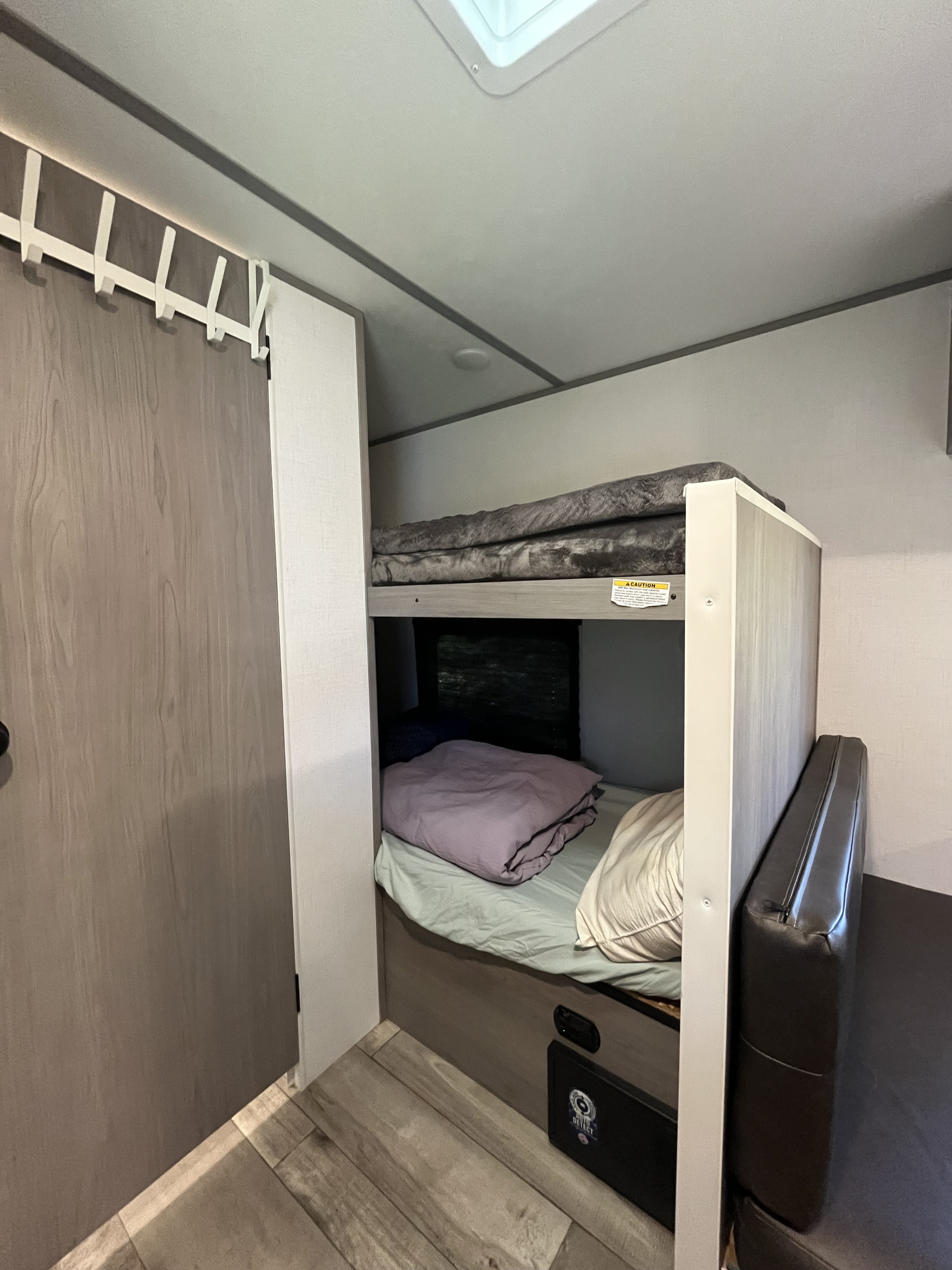 RV rental interior: stacked bunk beds with gray and purple comforters, a window, and a dark brown upholstered seat.. Coachmen 17b 2024
