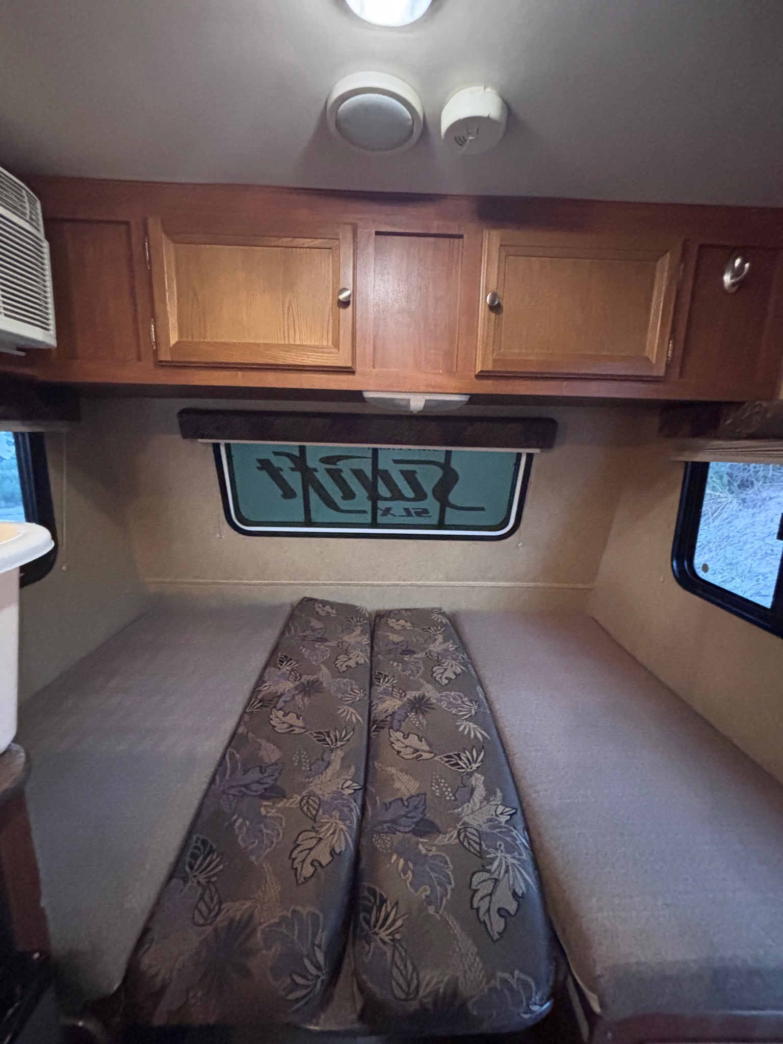 Cozy RV rental interior features a sleeping area with patterned cushions, overhead cabinets, and windows. Includes AC and smoke detector.. Jayco Jay Flight Swift SLX 2013