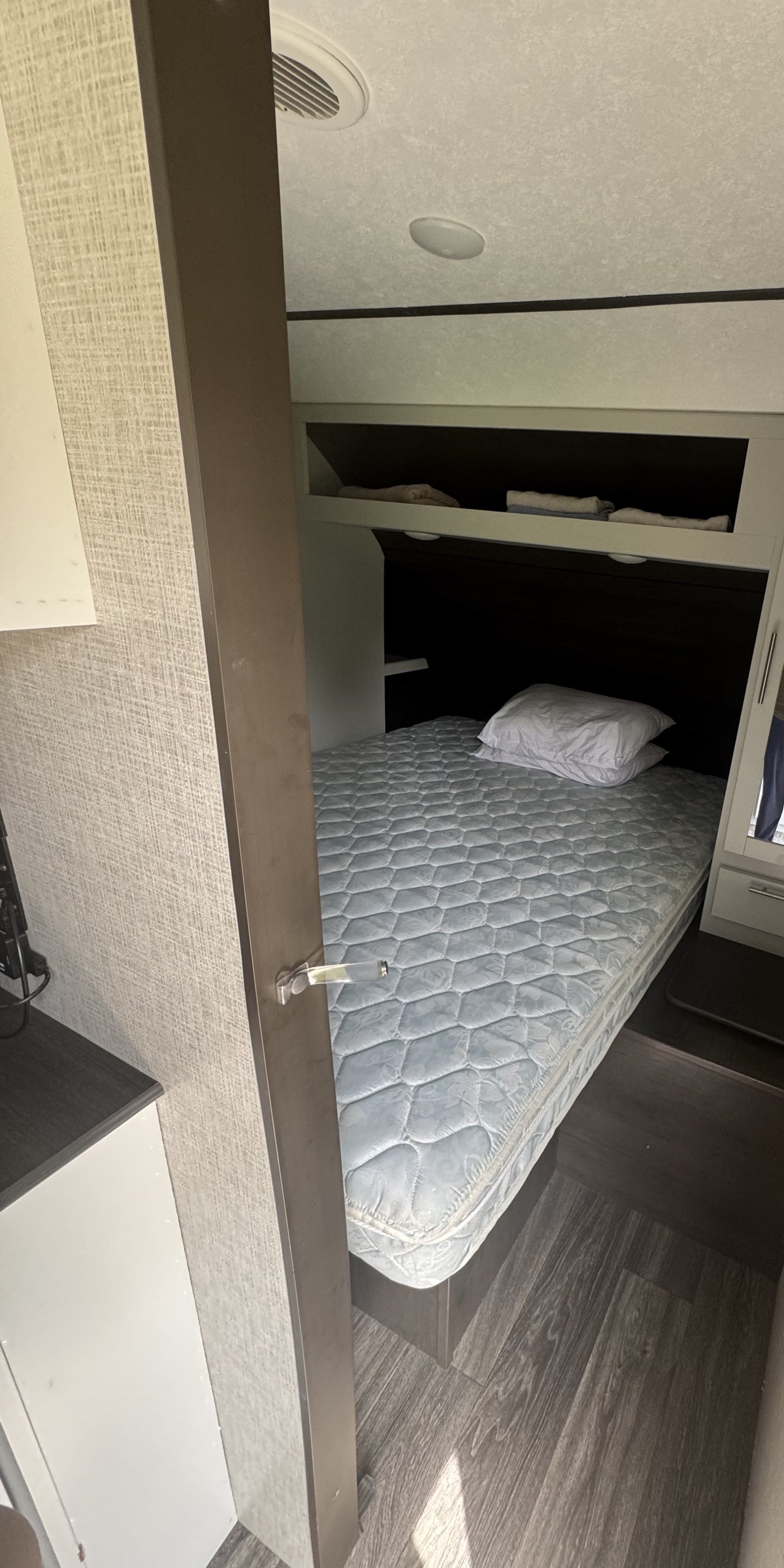 View into a compact RV rental bedroom with a patterned mattress, white pillow, and overhead shelves. Textured walls, wood-look floor.. Keystone RV Bullet Ultra Lite 2021