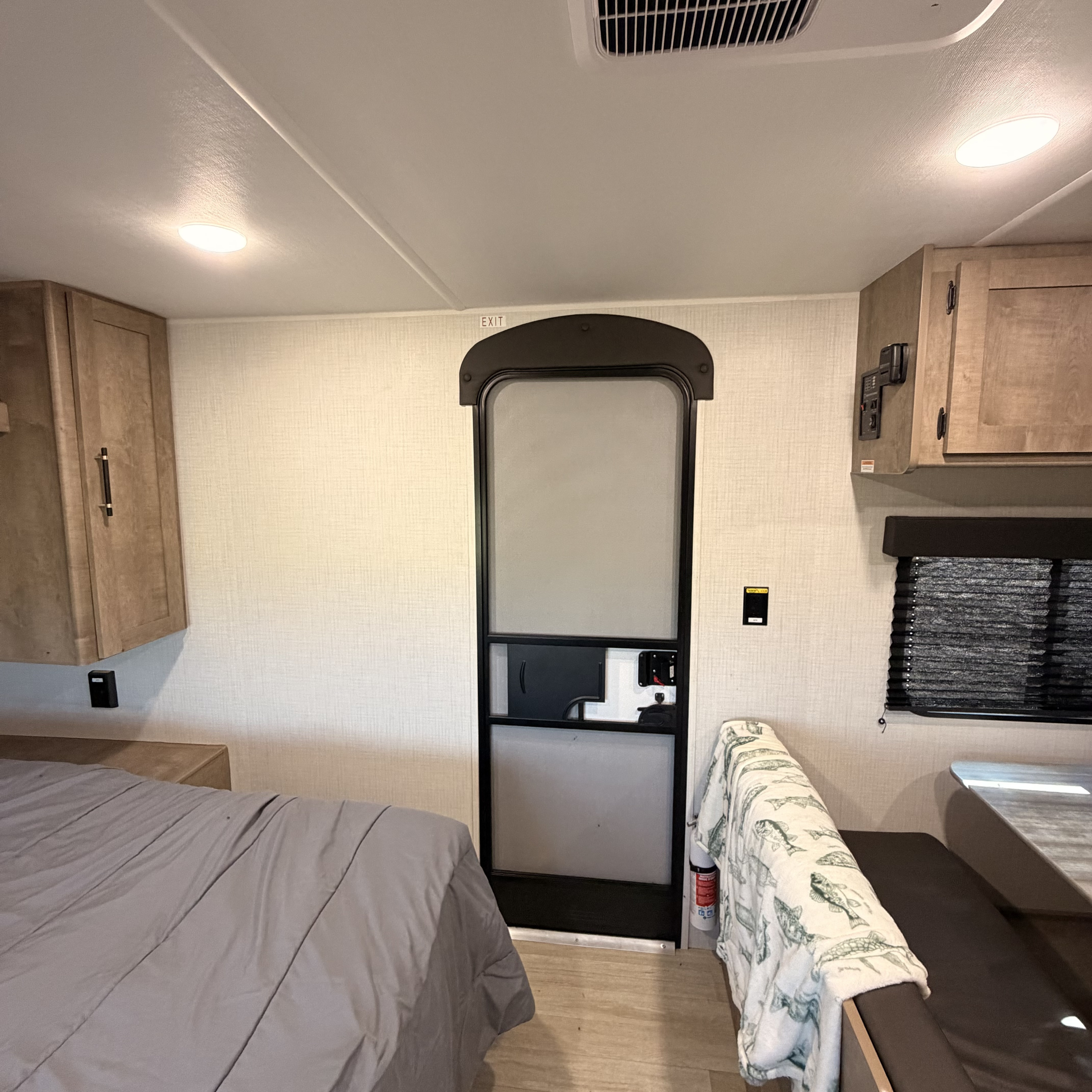 Interior of an RV rental: a bed, central door (with an 'EXIT' sign), cabinets, and a dinette with a fish blanket.. Keystone RV Springdale Mini 2026