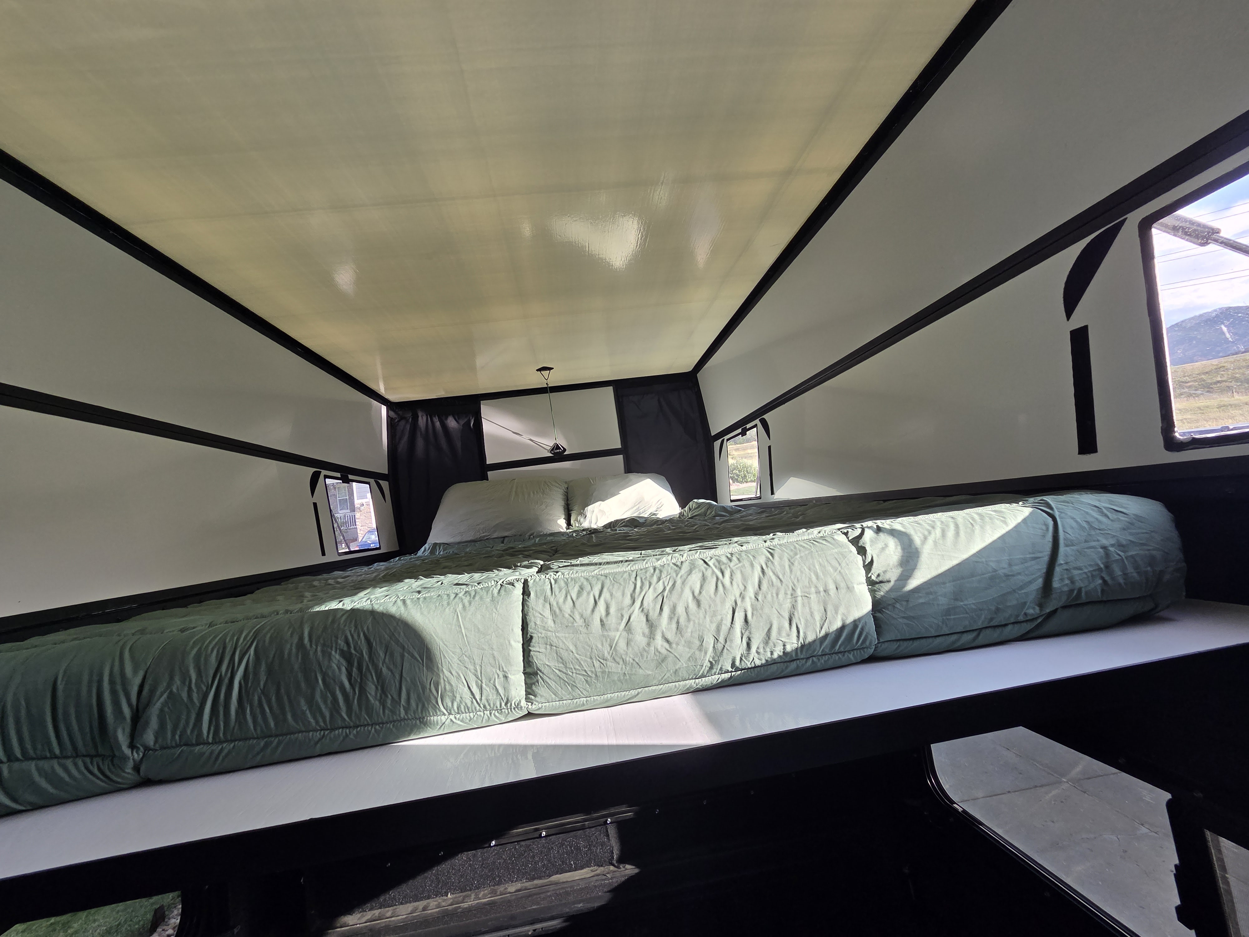 Interior view of an RV rental sleeping area, showing a large bed with green bedding, white walls, and a light pop-up roof.. Jeep Gladiator 2020