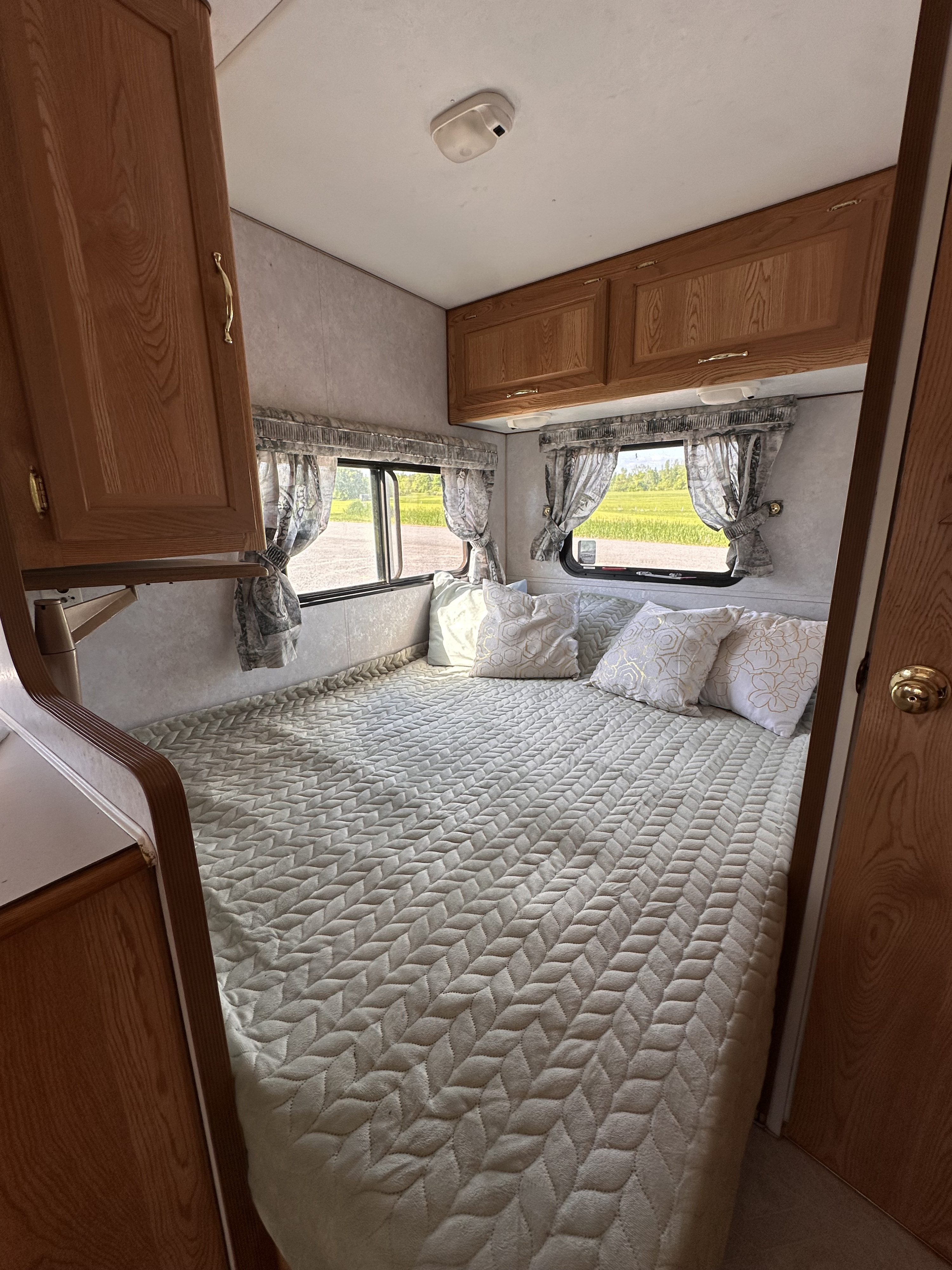 An RV rental bedroom with a comfy quilted bed, decorative pillows, wooden cabinets, and sunny windows overlooking a field.. Sun-Lite Sun-Lite Trailer 2000