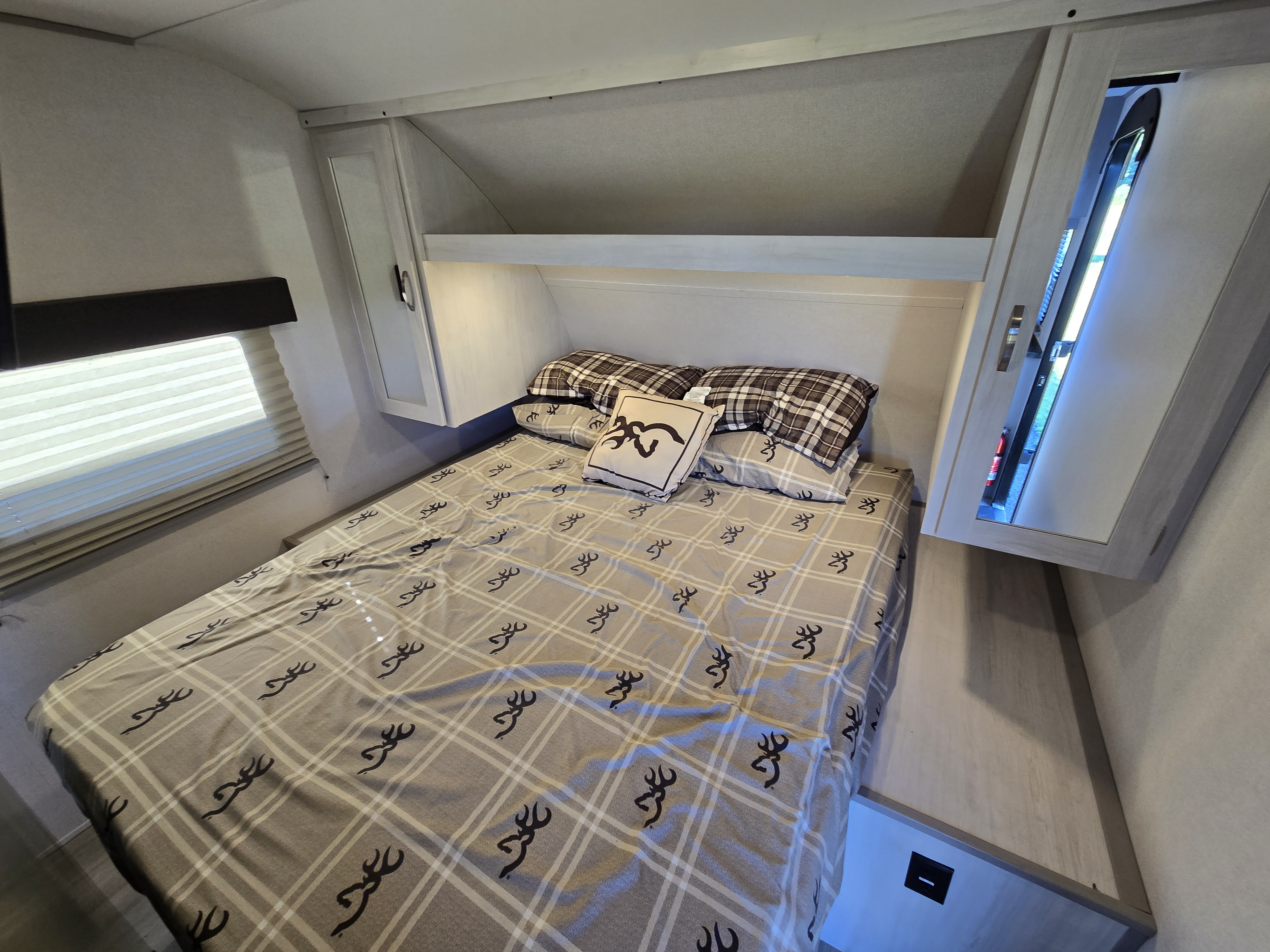 Cozy RV rental bedroom featuring a bed with plaid and deer logo bedding, overhead storage, and a window with blinds.. Forest River Tracer 2022