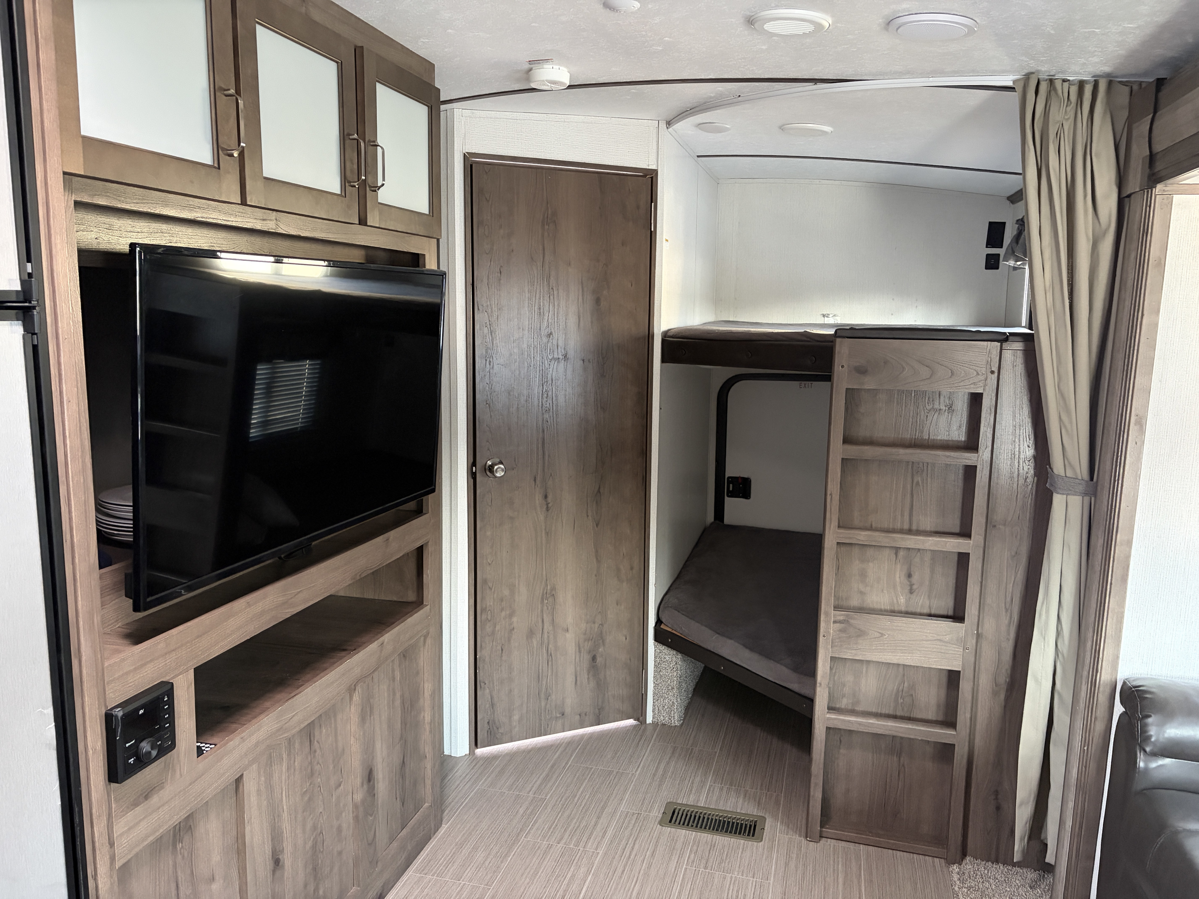 Inside an RV rental: view of wooden bunk beds with ladder, TV, upper cabinets, and a closed door, ideal for a comfortable stay.. Keystone RV Passport GT 2021