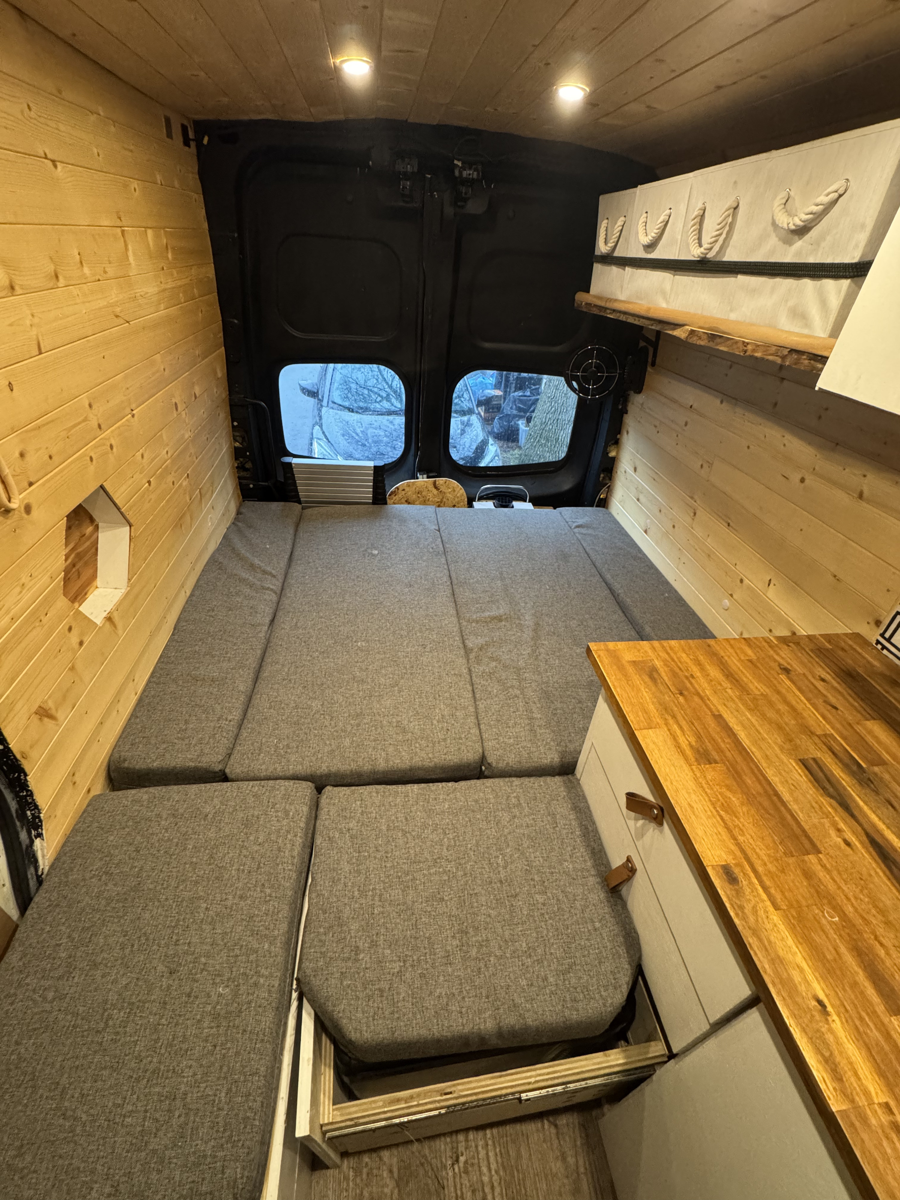 Cozy RV rental interior: wooden walls, comfy grey bed cushions, compact kitchen counter, and ample storage.. Nissan NV3500 2012
