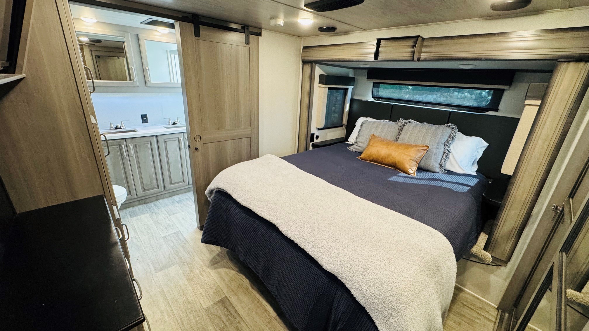 Inside this modern RV rental: a cozy bedroom with a dark bed, soft blankets, and an ensuite bathroom with dual sinks.. Keystone RV Montana 2021
