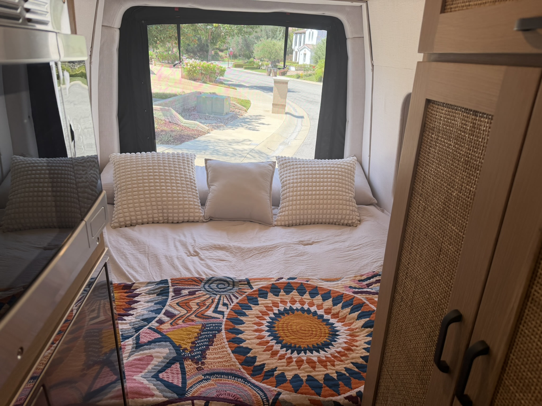 Cozy RV rental bed featuring a colorful quilt, white pillows, and a scenic view of a residential street through the rear window.. Mercedes-Benz sprinter 2025