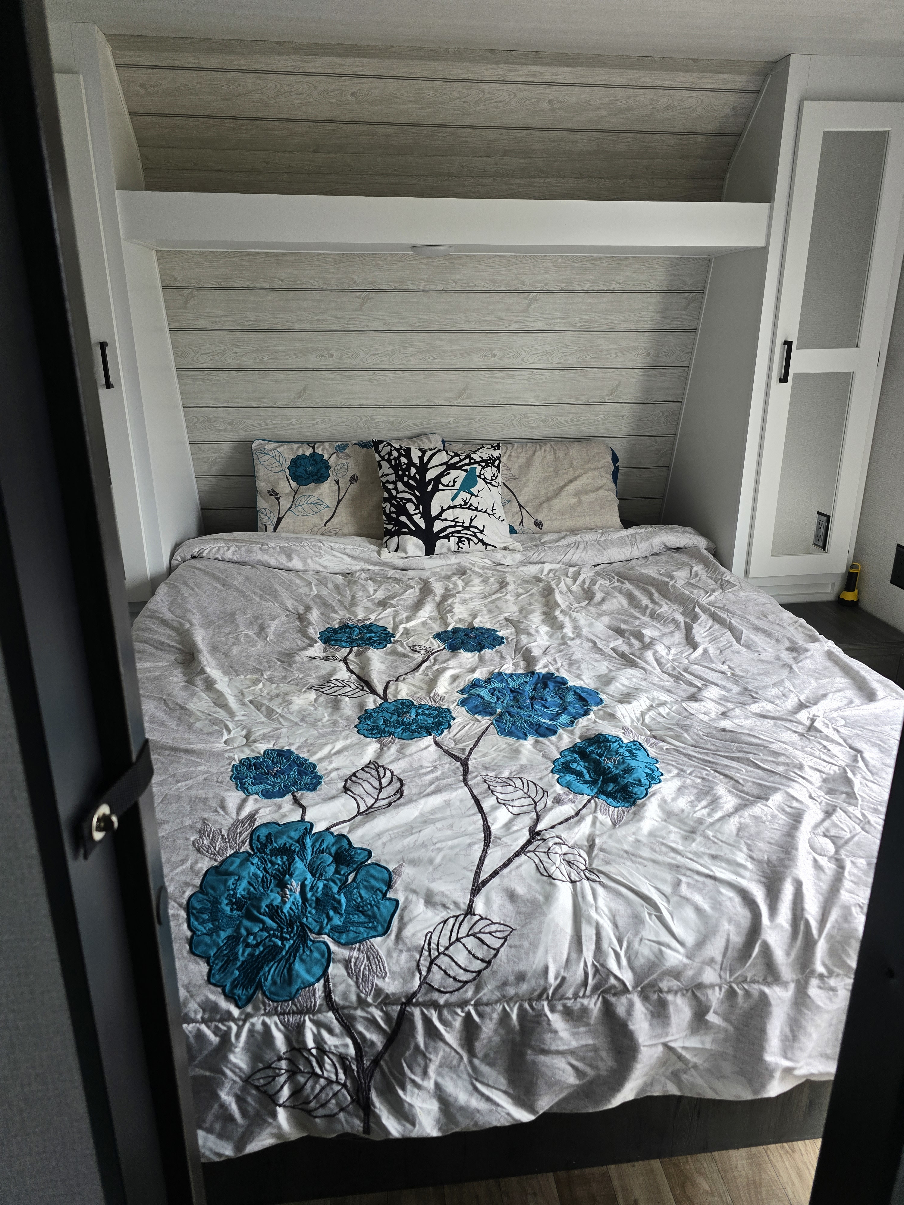 Relaxing RV rental bedroom. Features a comfortable bed with teal floral decor, wood-paneling, and built-in storage.. Keystone RV Hideout 2024
