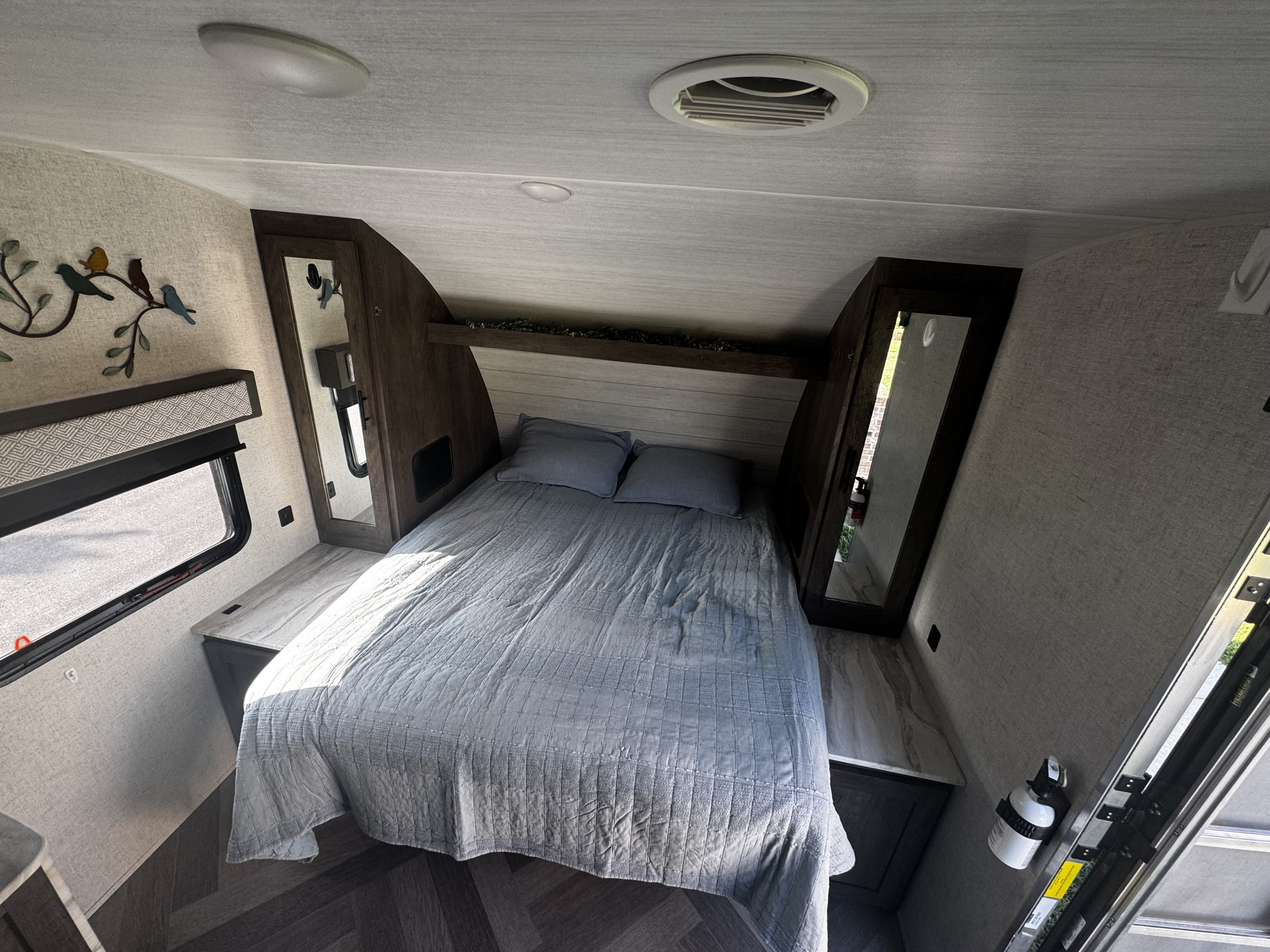 RV rental bedroom with a gray quilted bed, mirrored wardrobes, window, overhead shelf, ceiling lighting, and air vent.. Forest River Wildwood X-Lite 2021