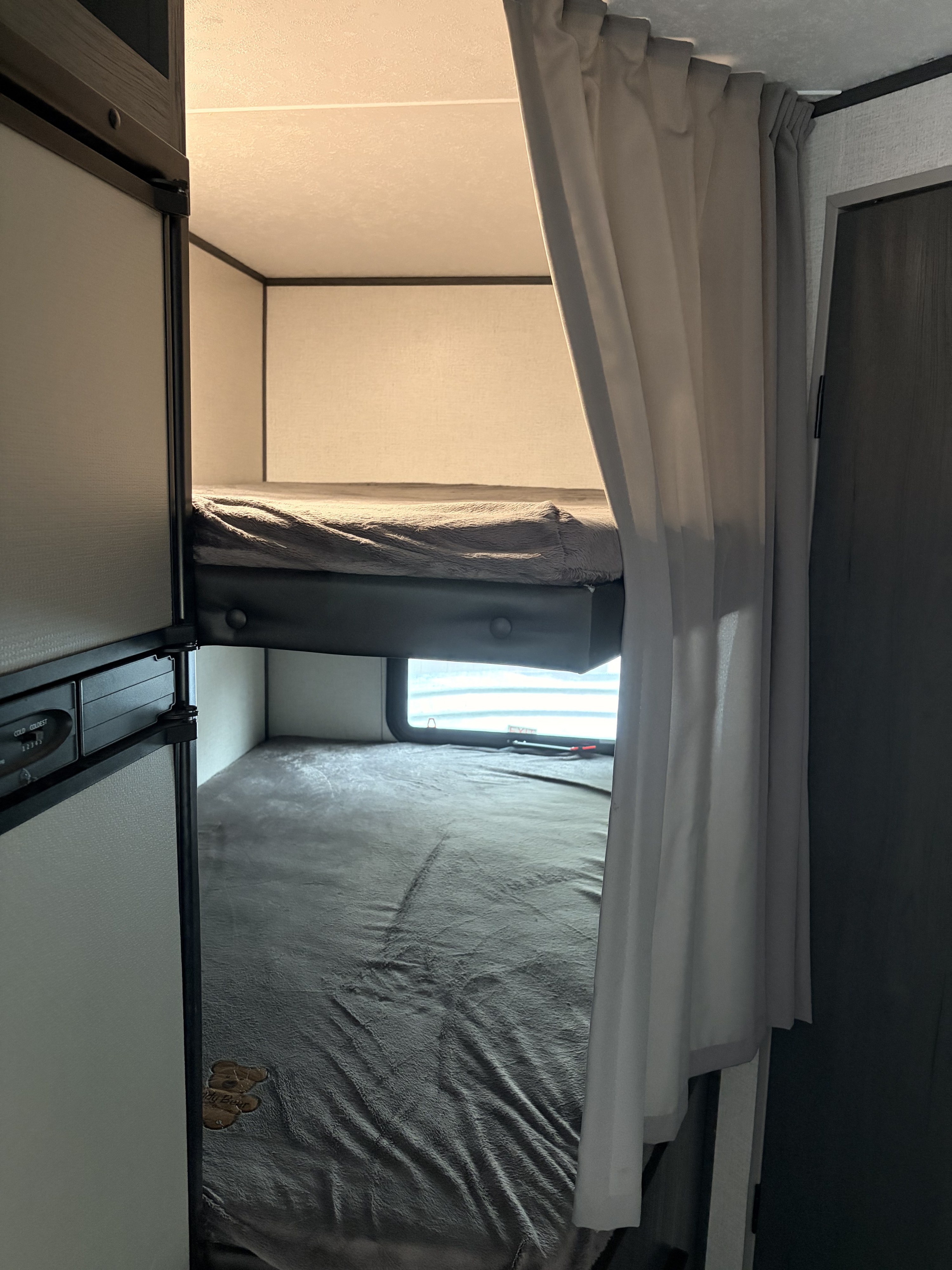 Inside an RV rental: view of the bunk beds with grey bedding, a partial curtain, and a refrigerator to the left.. Aspen Trail Aspen Trail Trailer 2021