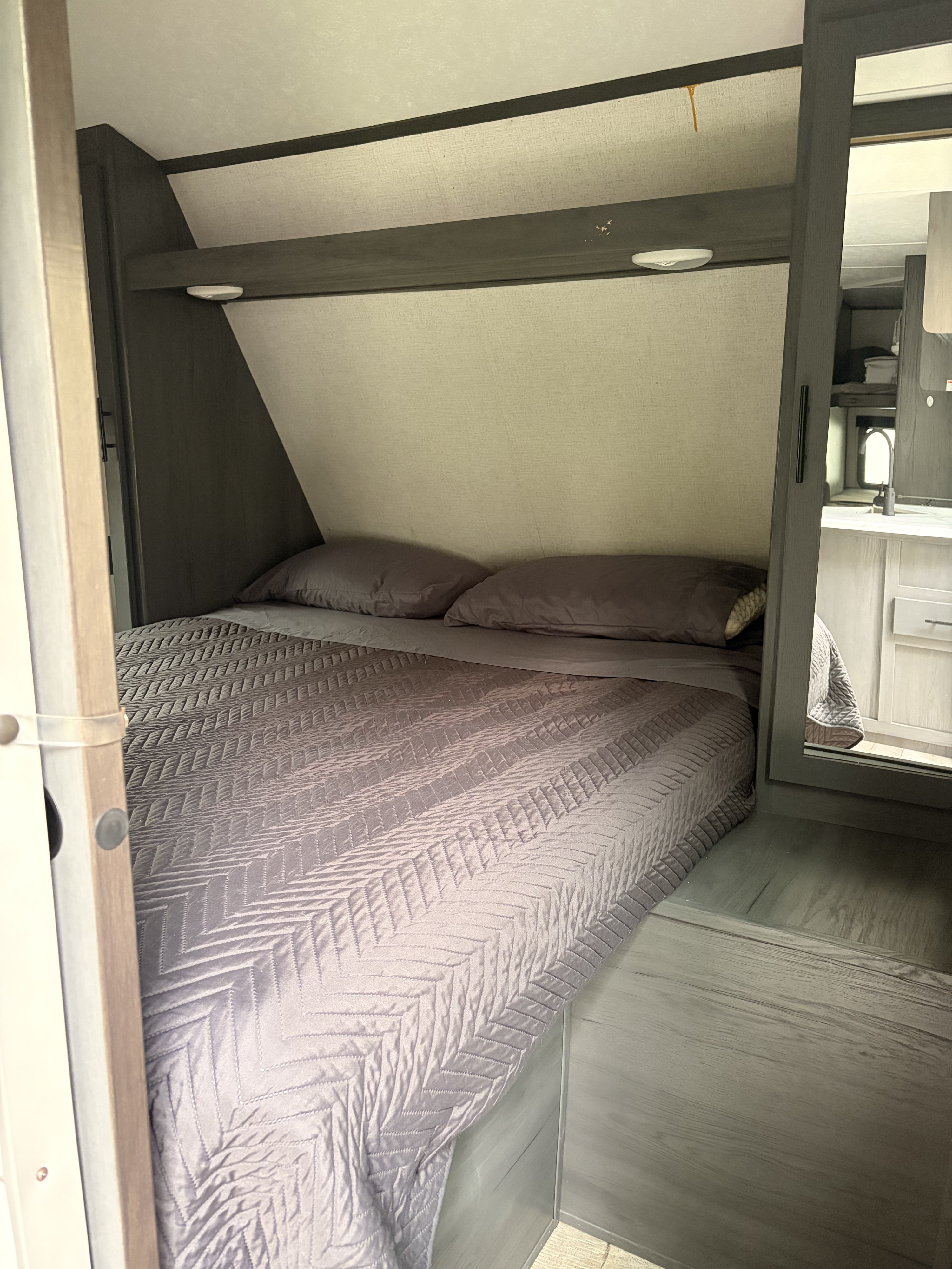 This RV rental's bedroom shows a cozy bed with gray bedding, an overhead shelf, and a reflected view of the kitchenette.. Aspen Trail Aspen Trail Trailer 2021