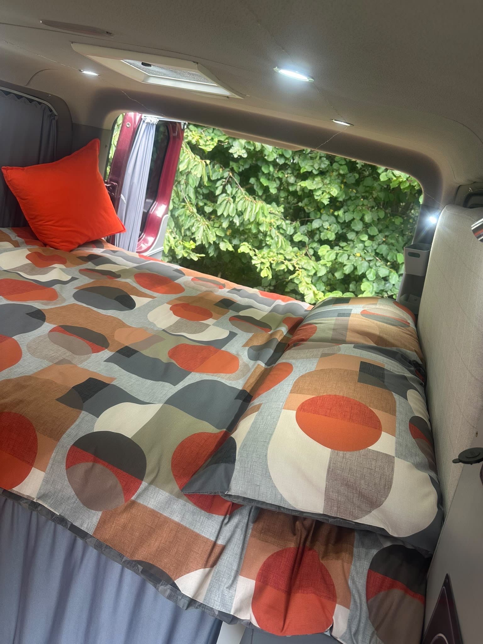 Experience van life! A colorful, comfy campervan bed in an RV rental with an orange pillow and bright interior lights, looking out to nature.. Mercedes Sprinter 2014