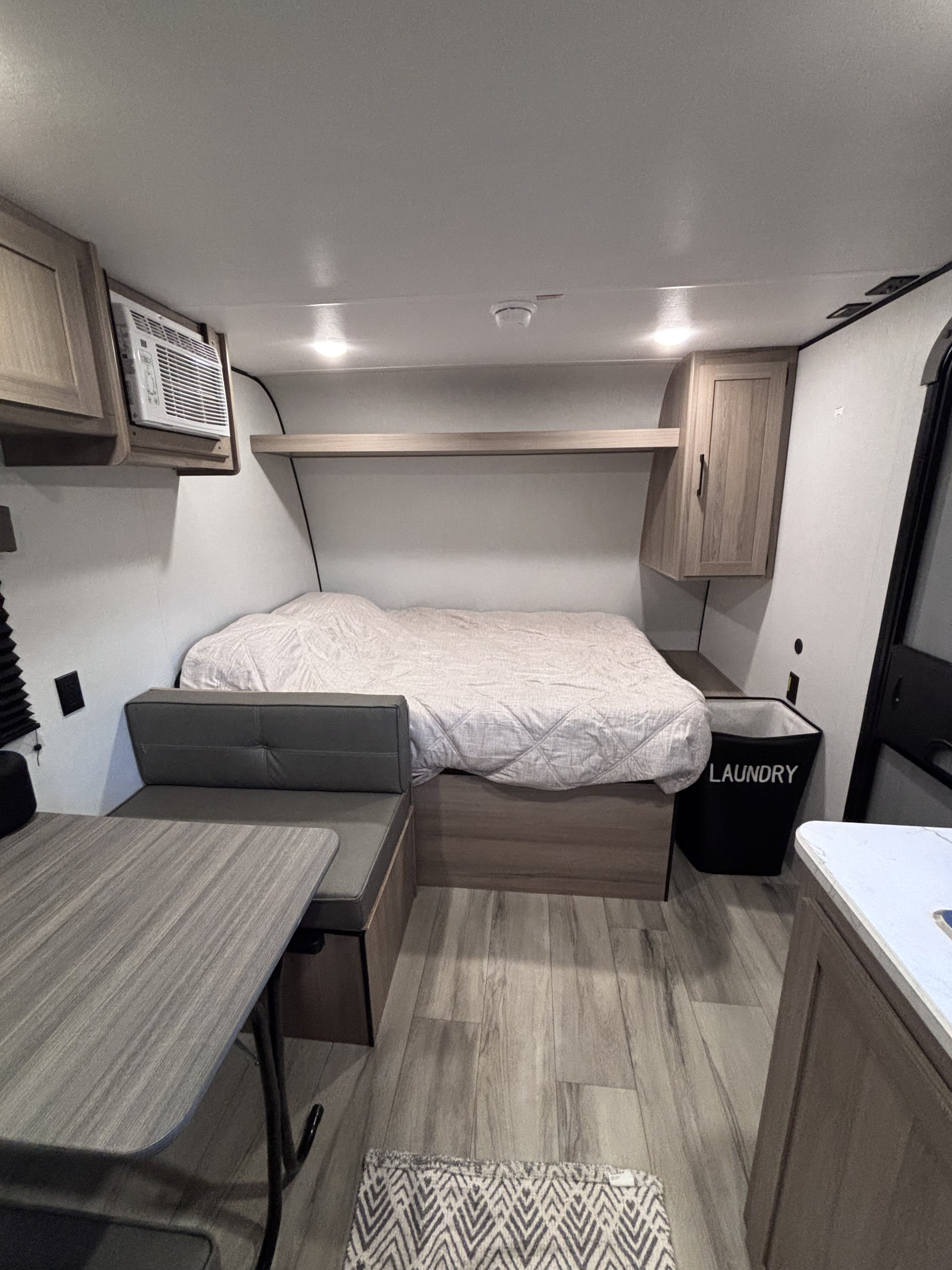 RV rental showing a cozy, compact interior with a bed, dining area, window AC, overhead storage, and a laundry hamper.. Keystone RV Coleman Light 2026