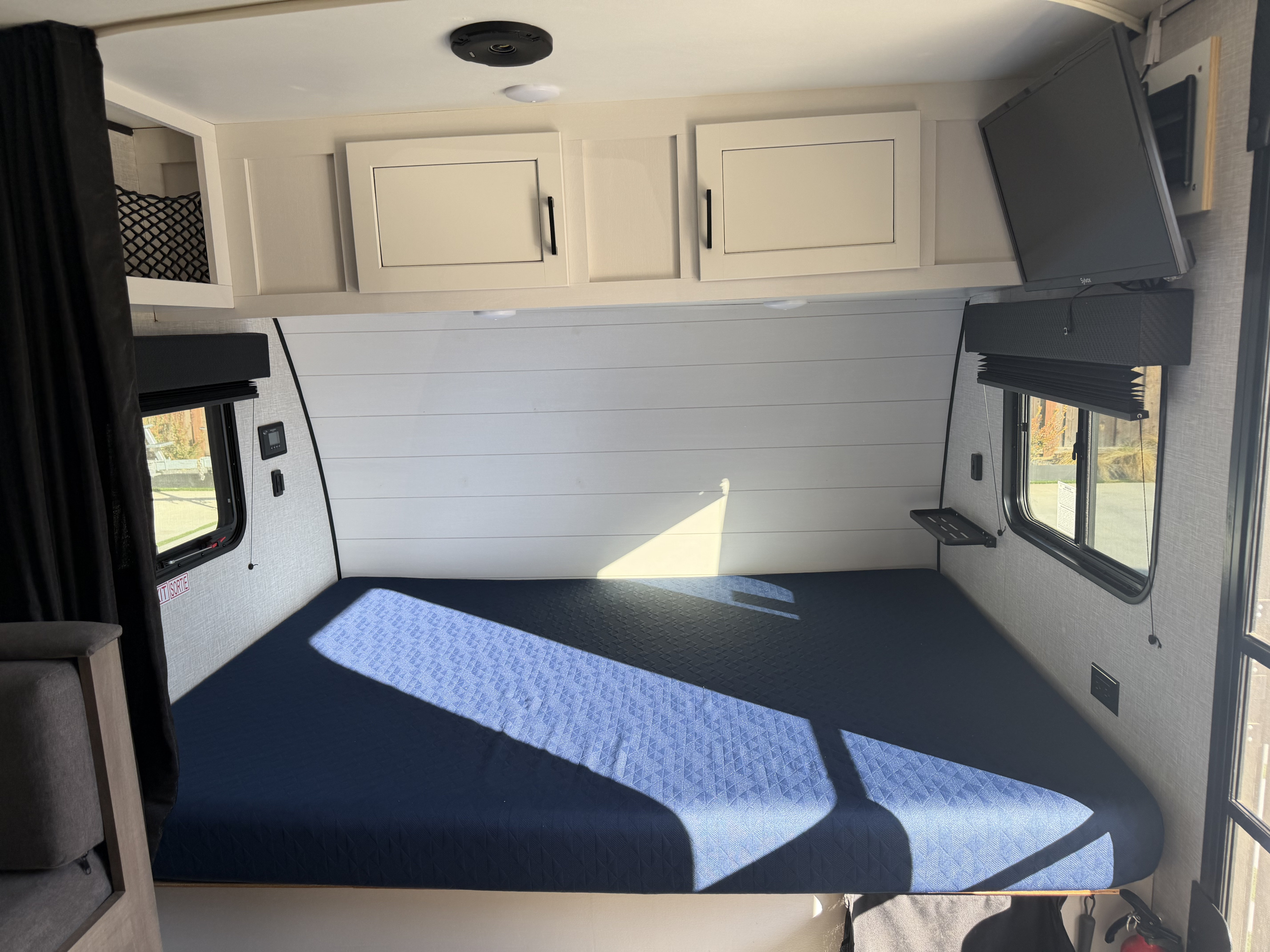 Interior of an **rv rental** featuring a compact bedroom with a blue bed, overhead cabinets, TV, and sunlit windows.. Jayco Jay Flight SLX Baja Edition 2024