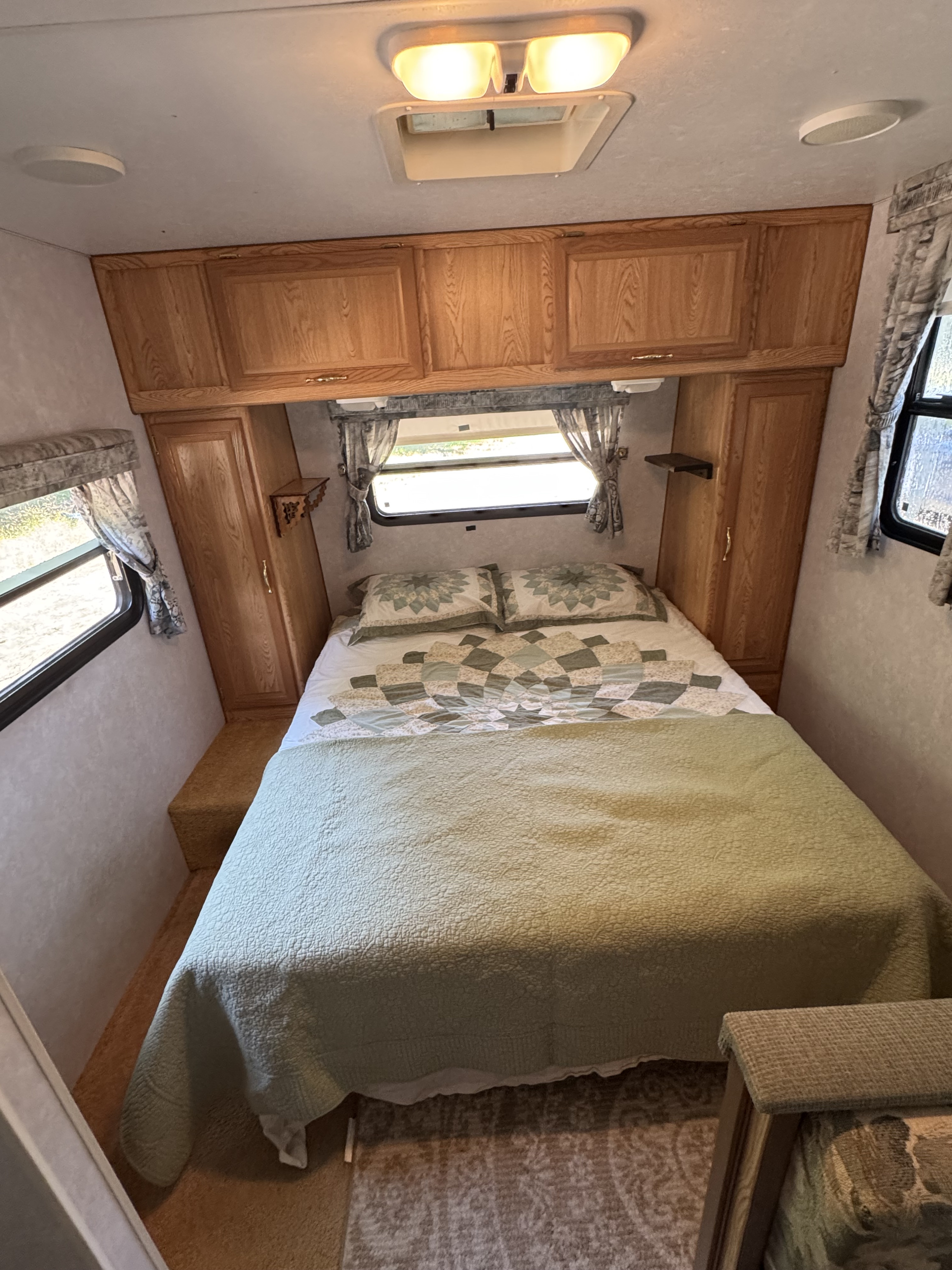 Inside an RV rental bedroom showing a bed with a green quilt, wood cabinets, and two windows.. Sunline Solaris Lite 2000