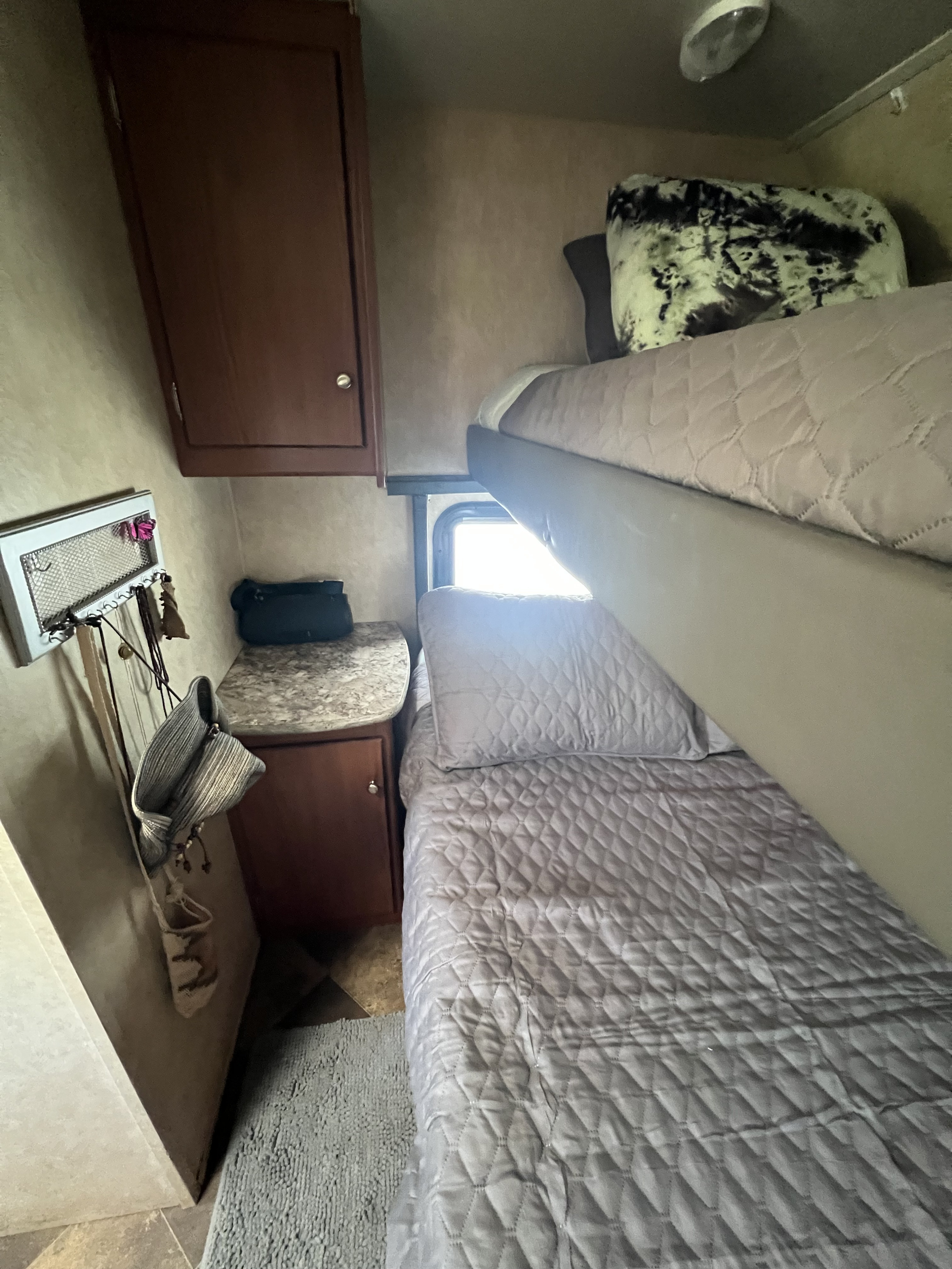 Cozy RV rental interior with bunk beds, quilted gray bedding, wooden cabinetry, and a bright window. Wall organizer visible.. Forest River Wildwood X-Lite FS 2015