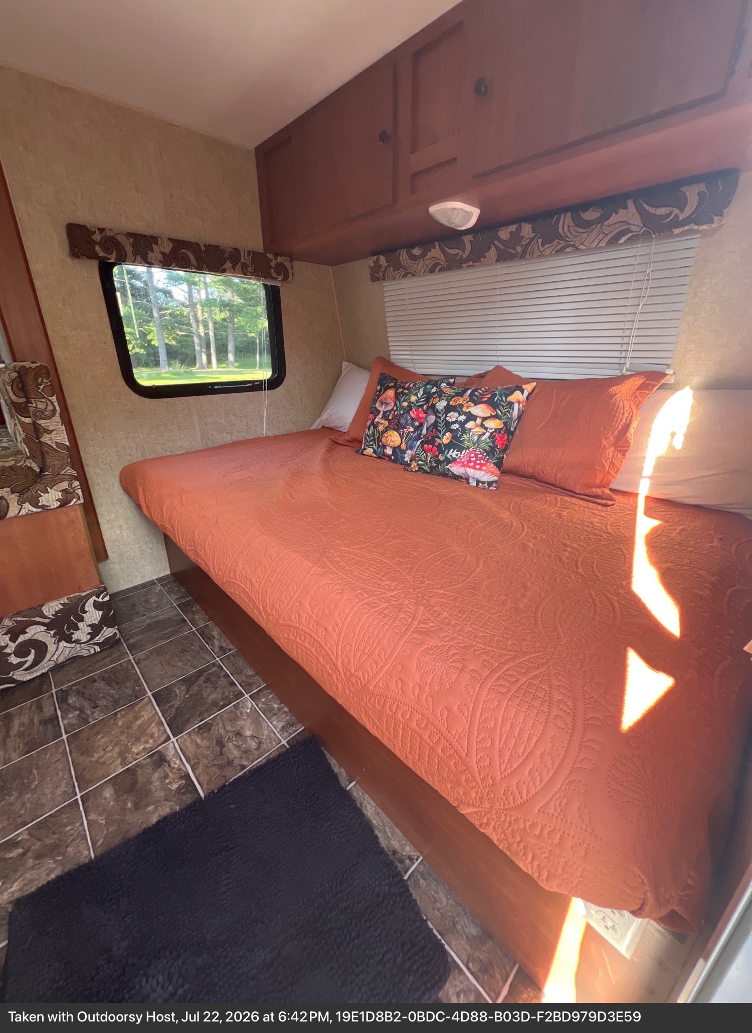 Cozy RV rental bedroom with an orange quilted bedspread, mushroom pillow, and a window view of trees. Ready for your next adventure!. Sportsmen Classic 2014