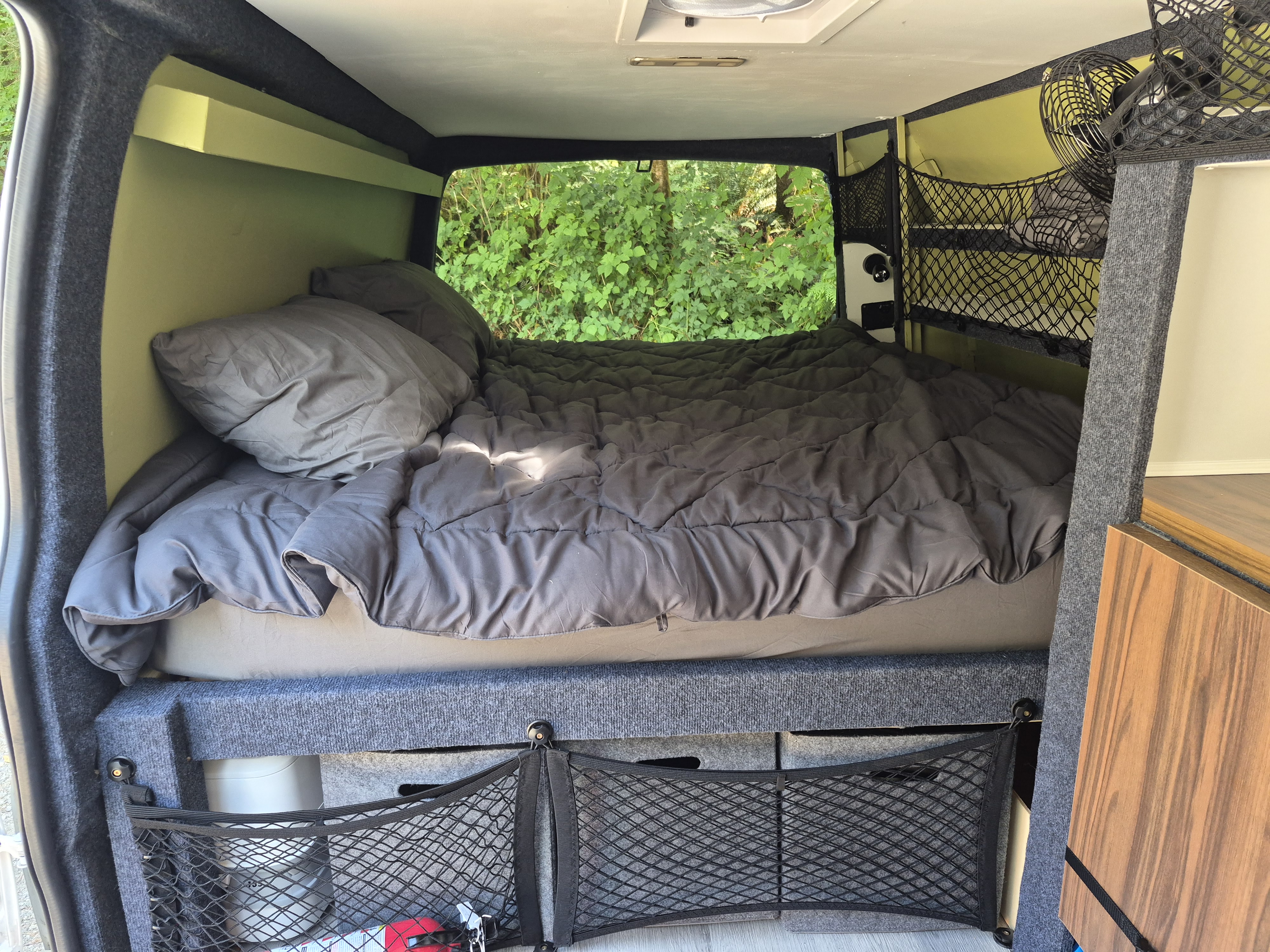 Comfortable bed in this RV rental's camper van interior. Green walls, lush nature view out back, & organized storage nets.. Chevrolet Express 2500 Extended 2015
