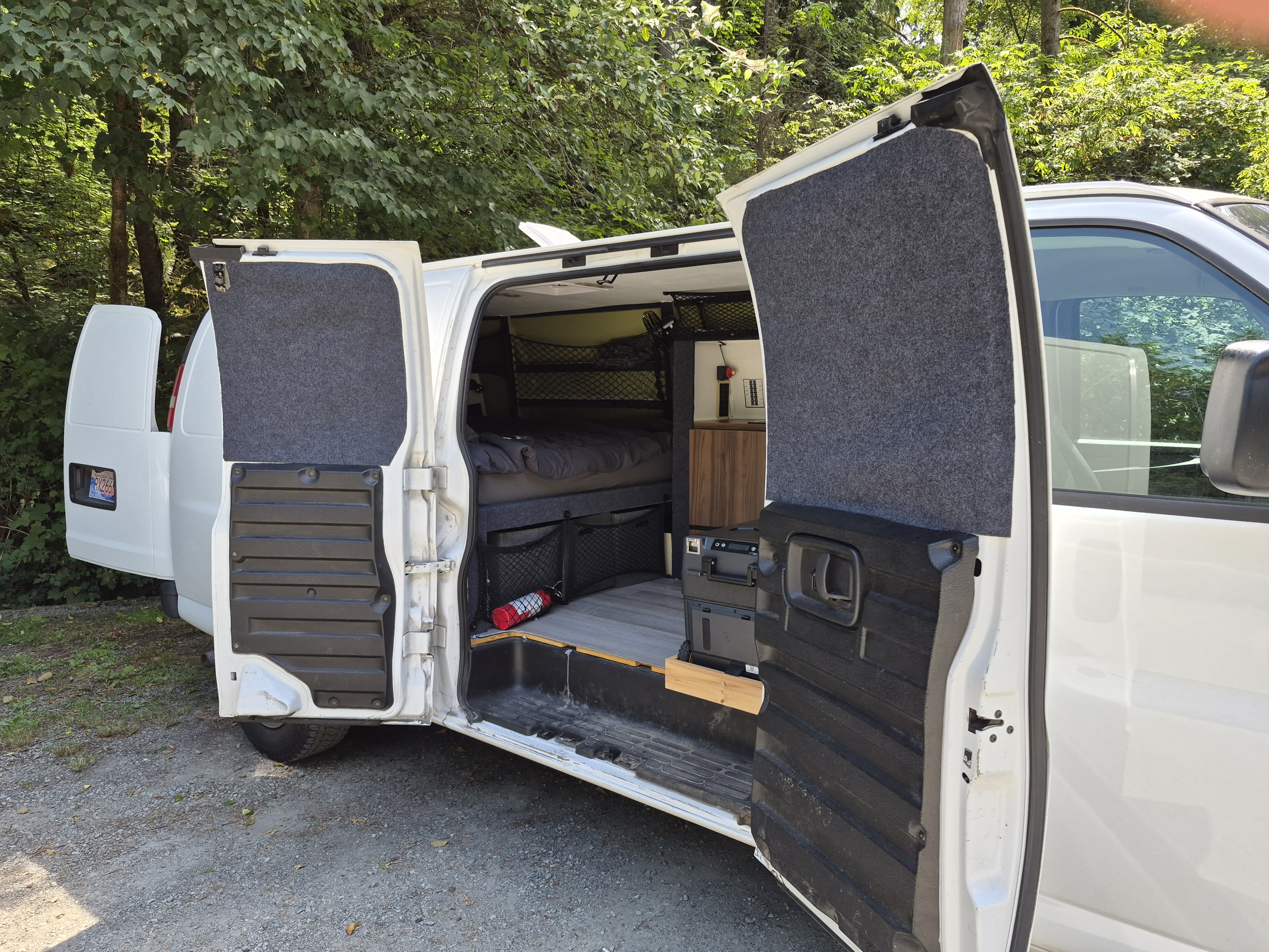 White RV rental campervan, side doors open to reveal its converted interior with a bed, storage nets, and portable fridge.. Chevrolet Express 2500 Extended 2015