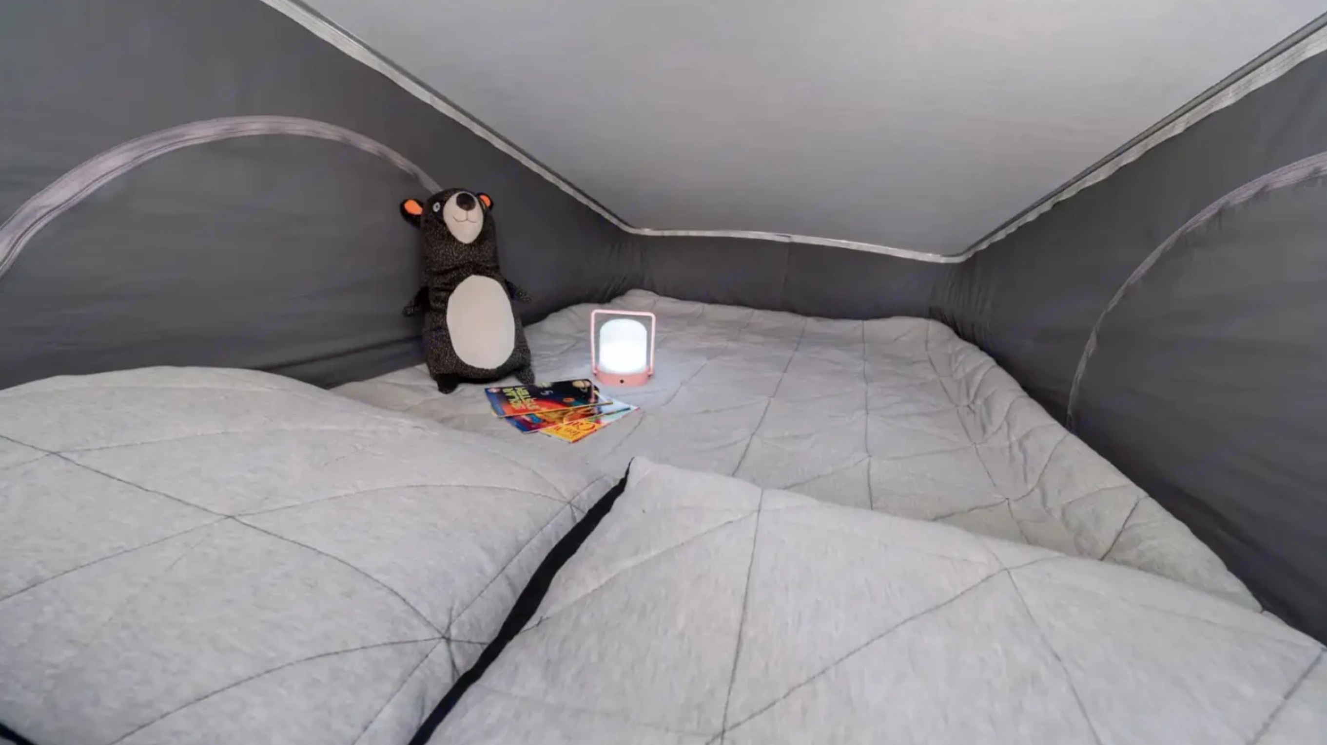 Inside a cozy RV rental: a grey quilted mattress, plush bear, books, and a glowing night light. Ideal for camper adventures.. Winnebago Solis 2023