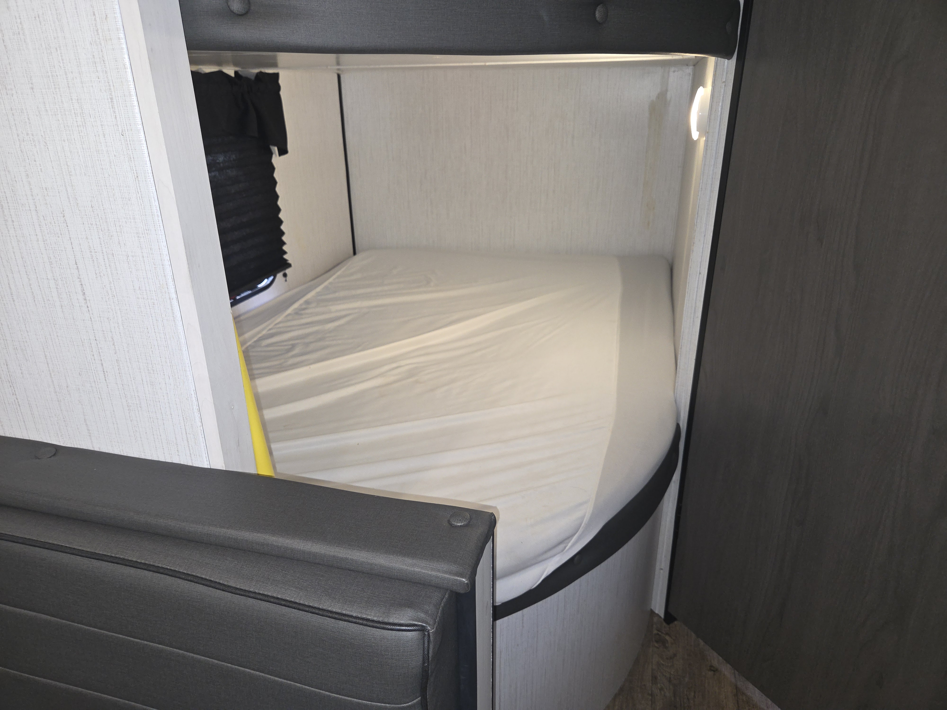 Inside an RV rental: a compact sleeping area with a bed, window with dark blinds, and a wall-mounted light.. Heartland Trail Runner 2022