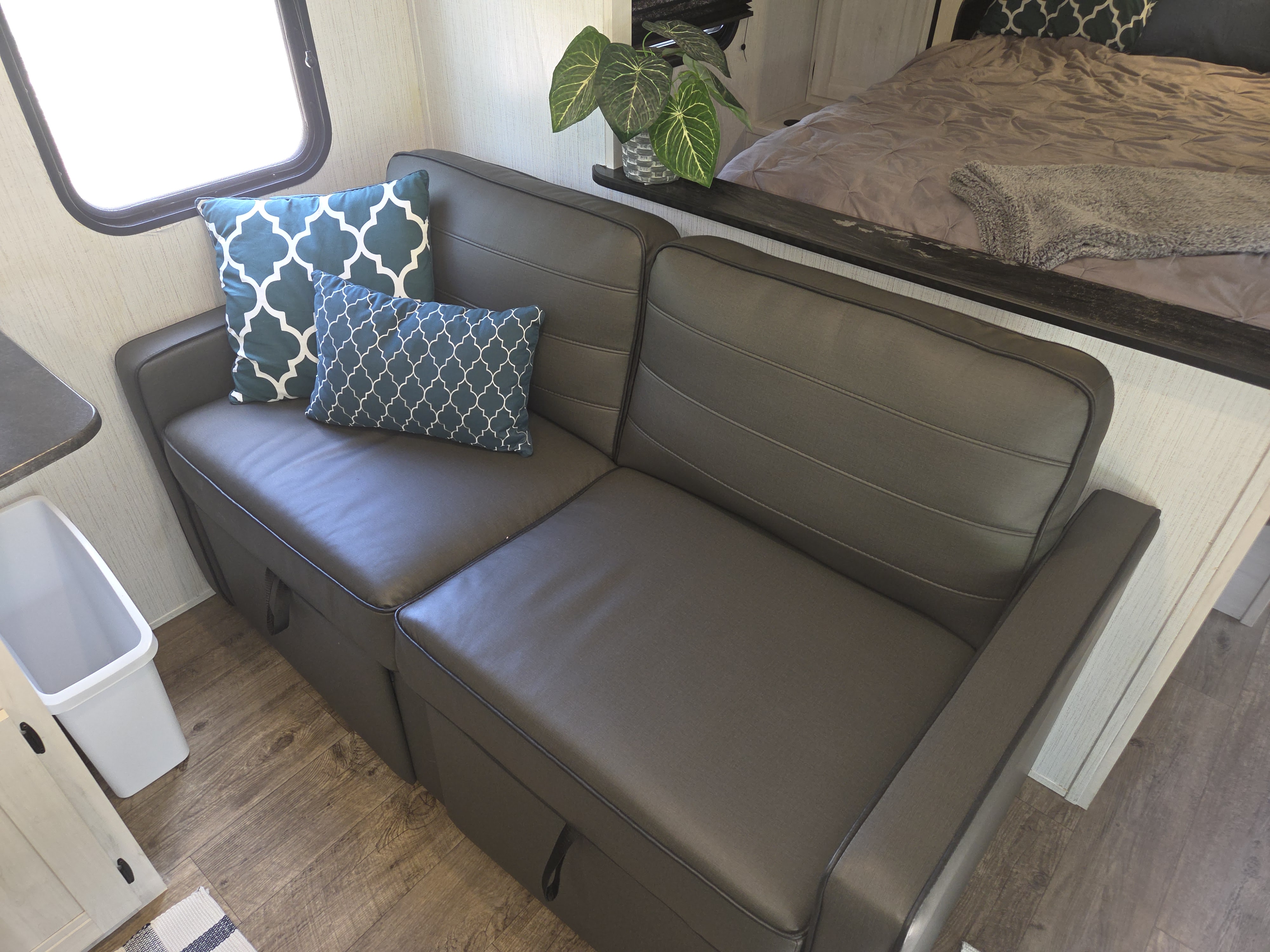 Cozy RV rental interior features a dark gray sofa with teal pillows, a plant, and a bed in a compact setting.. Heartland Trail Runner 2022