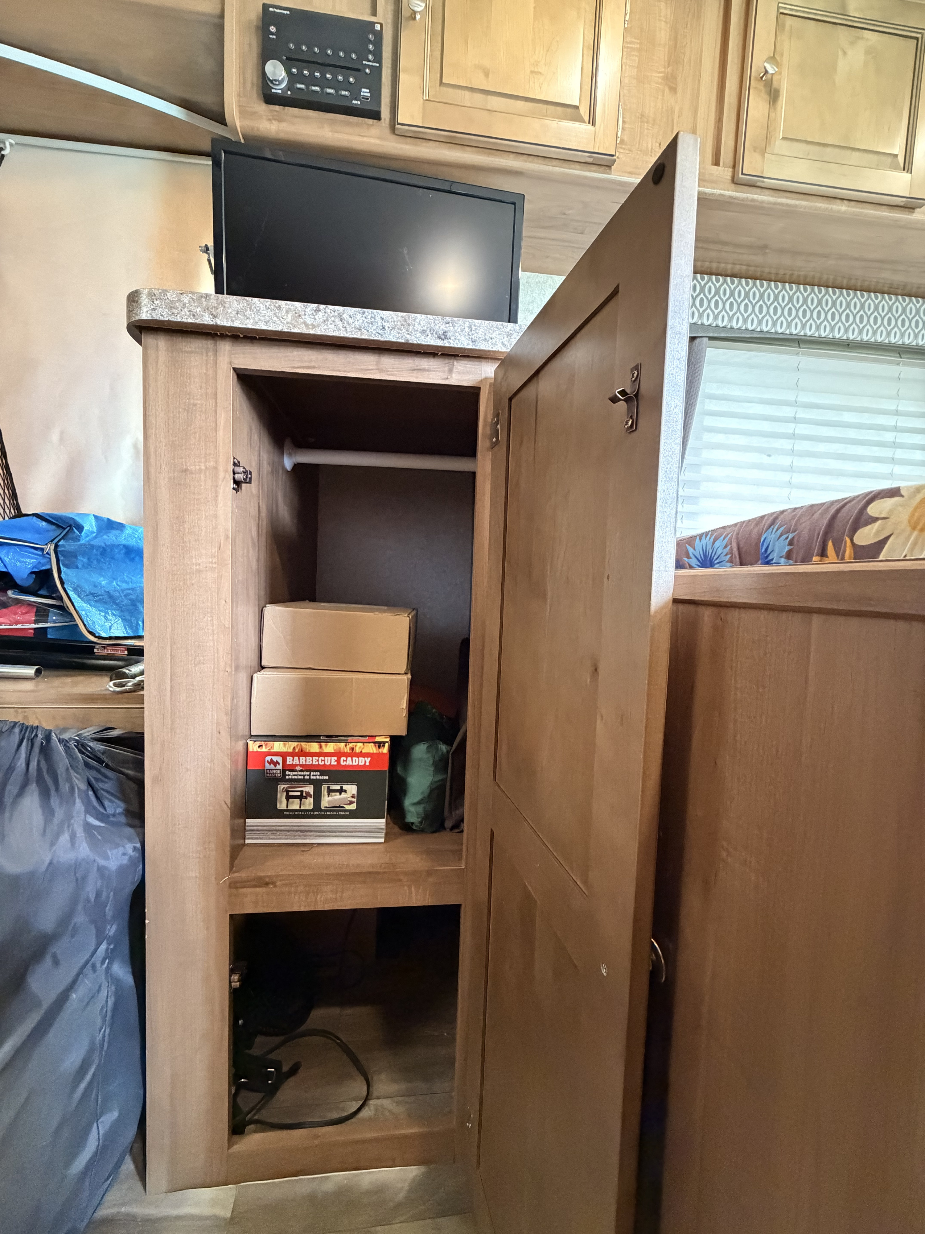 RV rental interior: An open wooden storage cabinet holds boxes and a barbecue caddy, with a TV and stereo above.. Forest River Flagstaff Shamrock 2018