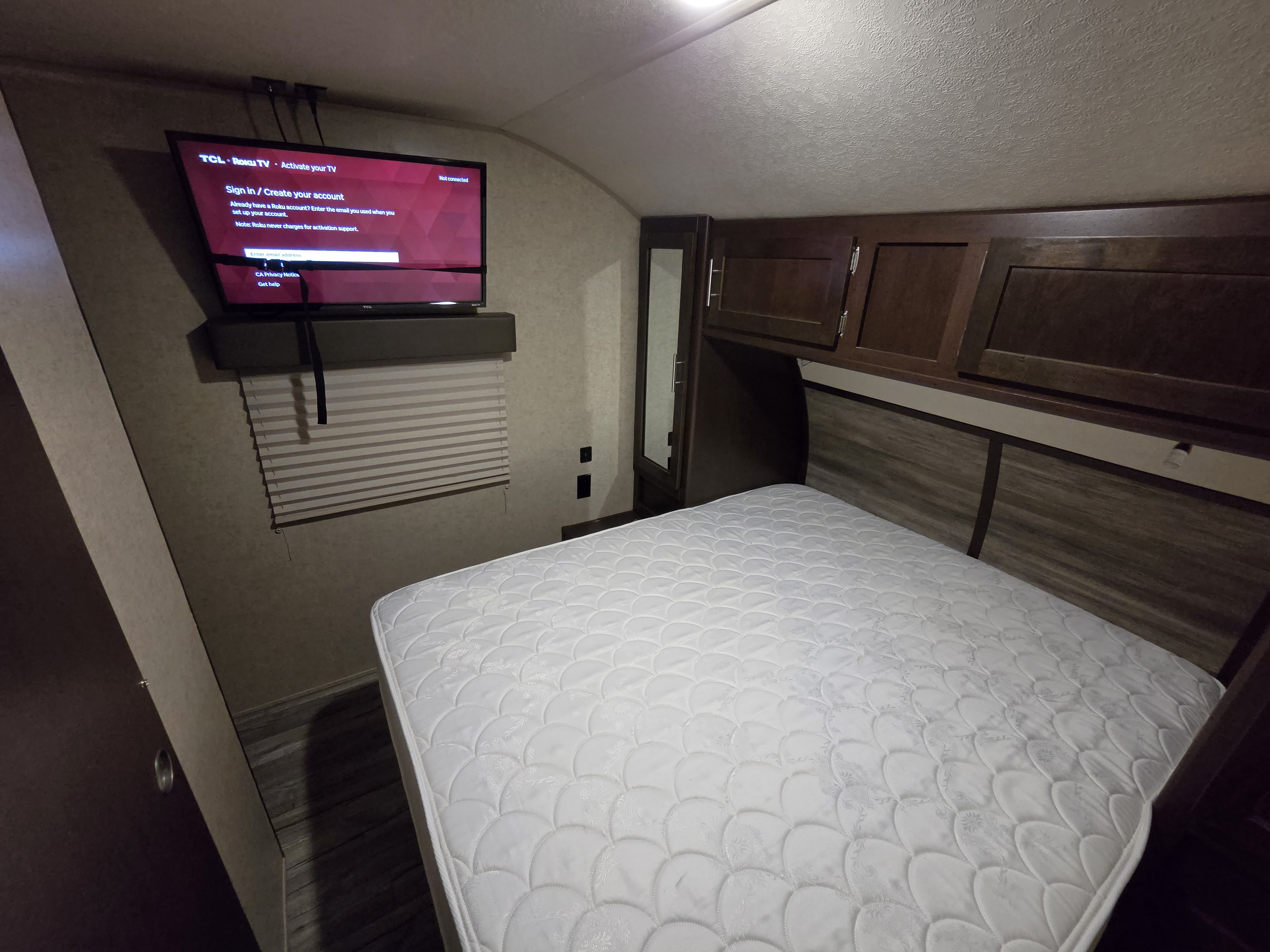 This RV rental bedroom features a comfortable white bed, overhead dark wood cabinets, and a TCL Roku TV displaying its activation screen.. Forest River Cherokee 2017