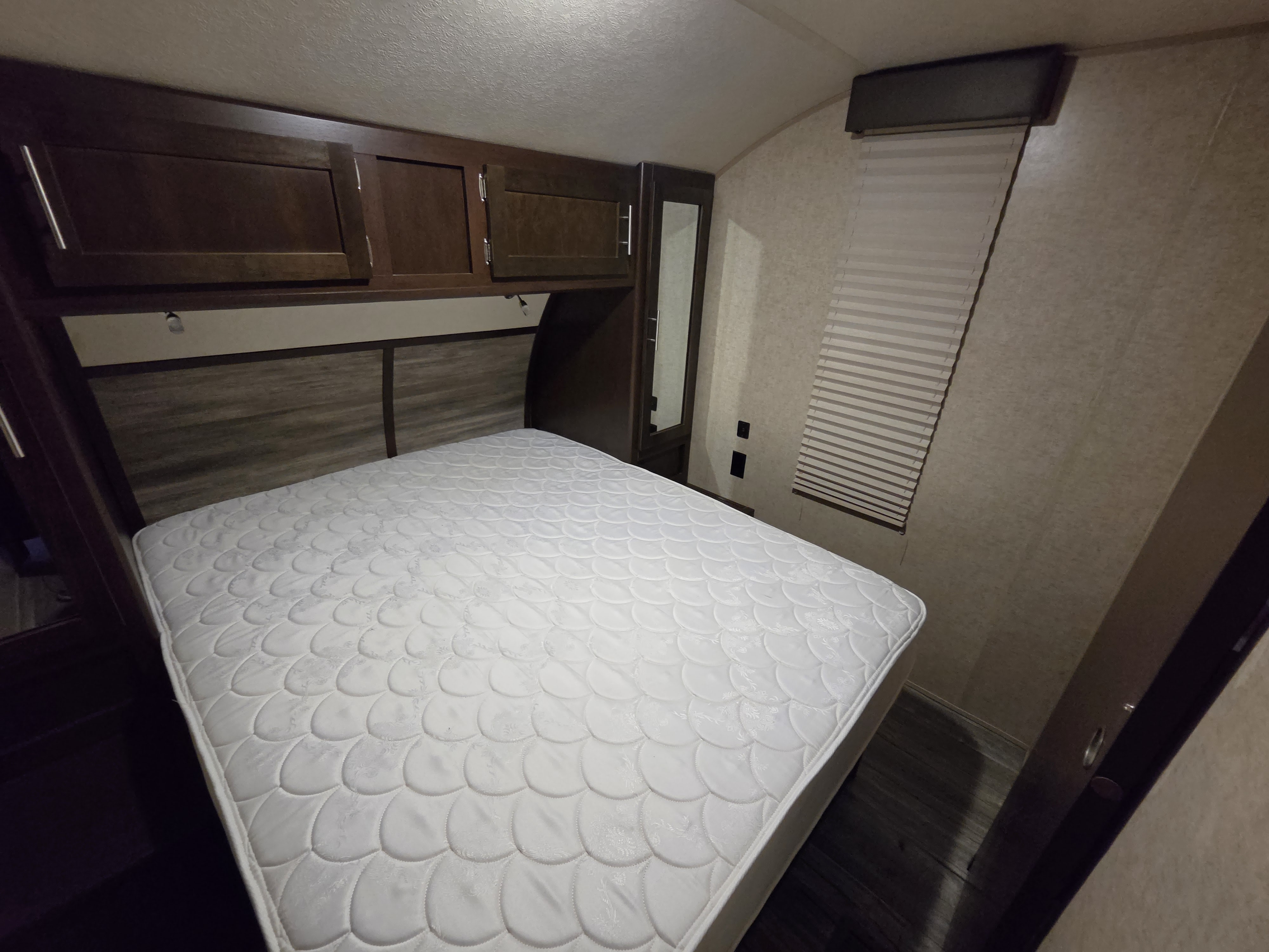 Inside an RV rental bedroom: a clean white mattress, dark wood overhead cabinets, wood headboard, and window blinds.. Forest River Cherokee 2017