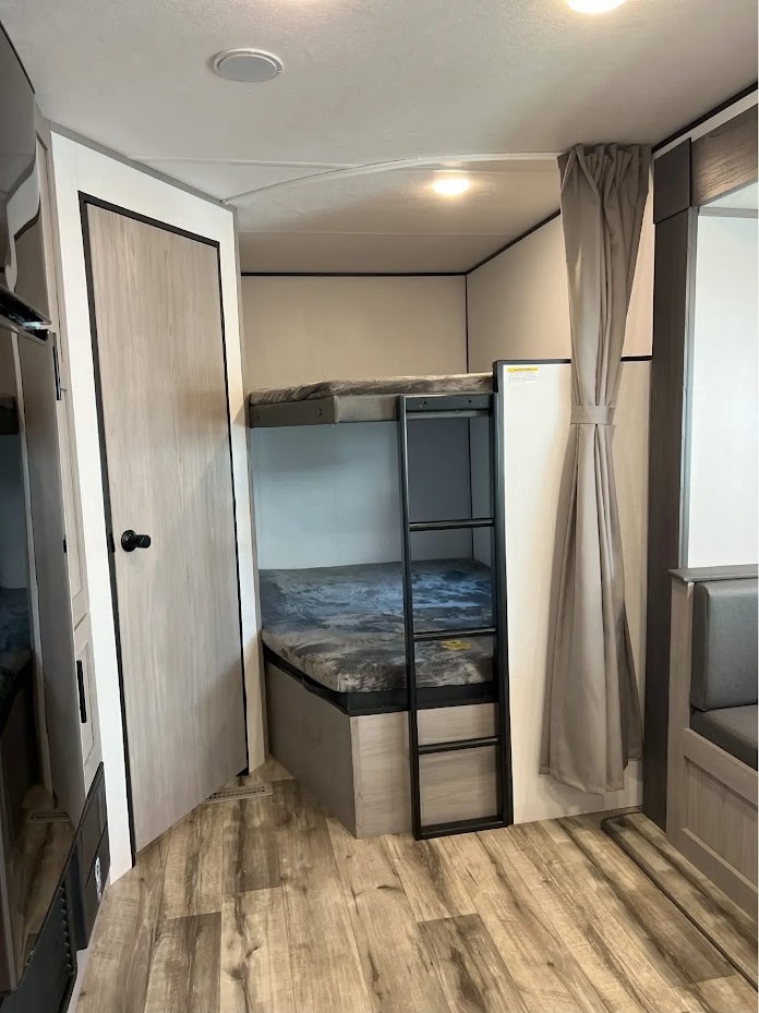 RV rental interior: Bunk beds with a ladder & privacy curtain. Wood flooring, door, and seating in this travel trailer space.. Dutchmen Coleman Lantern LT 2024
