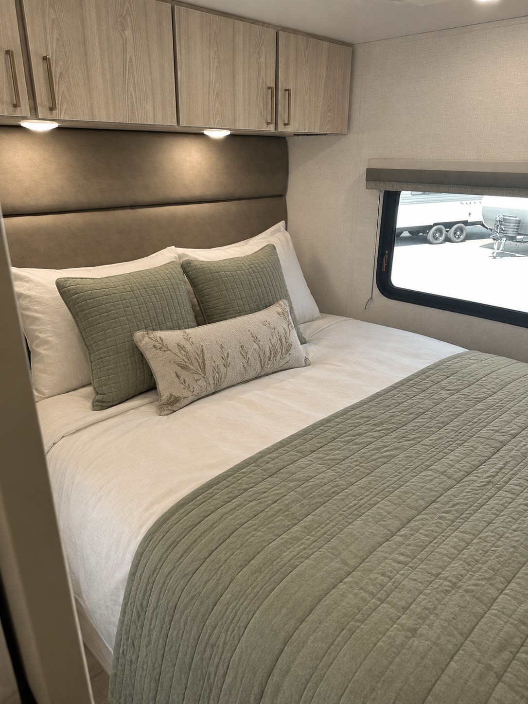Cozy RV rental bedroom featuring sage green bedding, padded headboard, overhead storage, and a window view.. East to West Entrada 2026